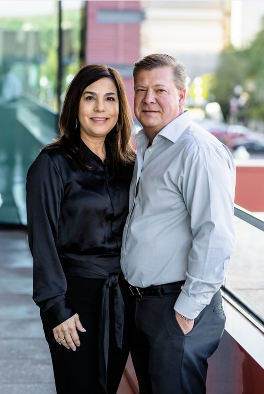 A middle-aged couple standing together in front of a large window, smiling at the camera. The woman has shoulder-length dark hair and is wearing a black satin long-sleeve blouse, while the man has short, gray hair and is dressed in a white button-up shirt and dark pants.
