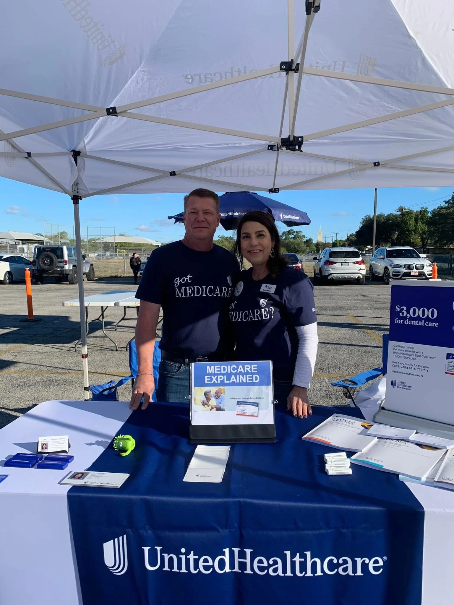 A woman and man standing behind a table at a United Healthcare booth, with a banner that says "Medicare Explained," under a white canopy tent, in an outdoor parking lot with parked cars and other booths visible in the background.