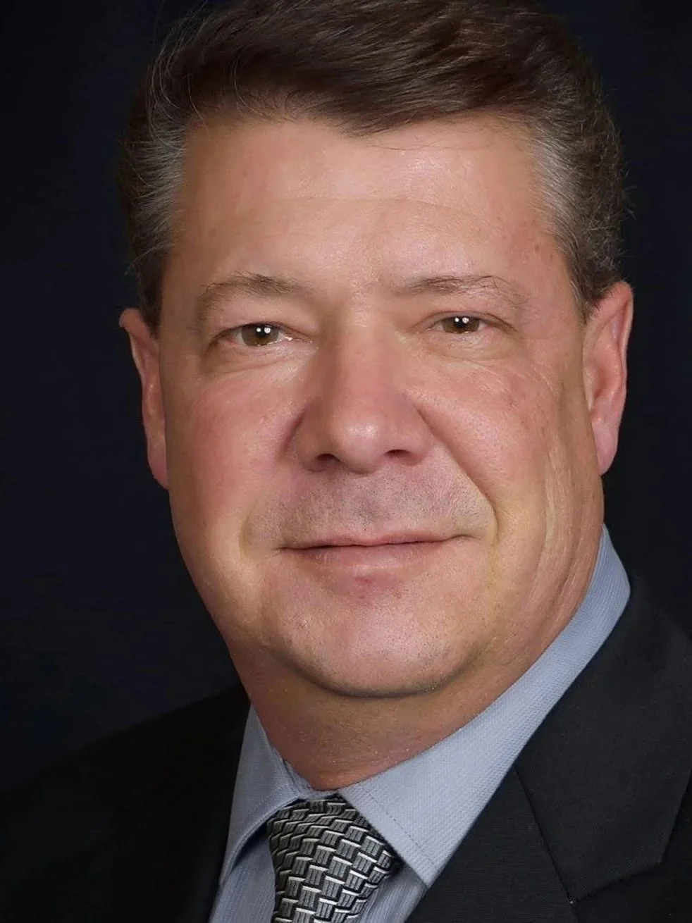 Close-up portrait of a middle-aged man with short, neatly combed brown hair, wearing a dark suit, light blue shirt, and patterned tie, against a dark background.