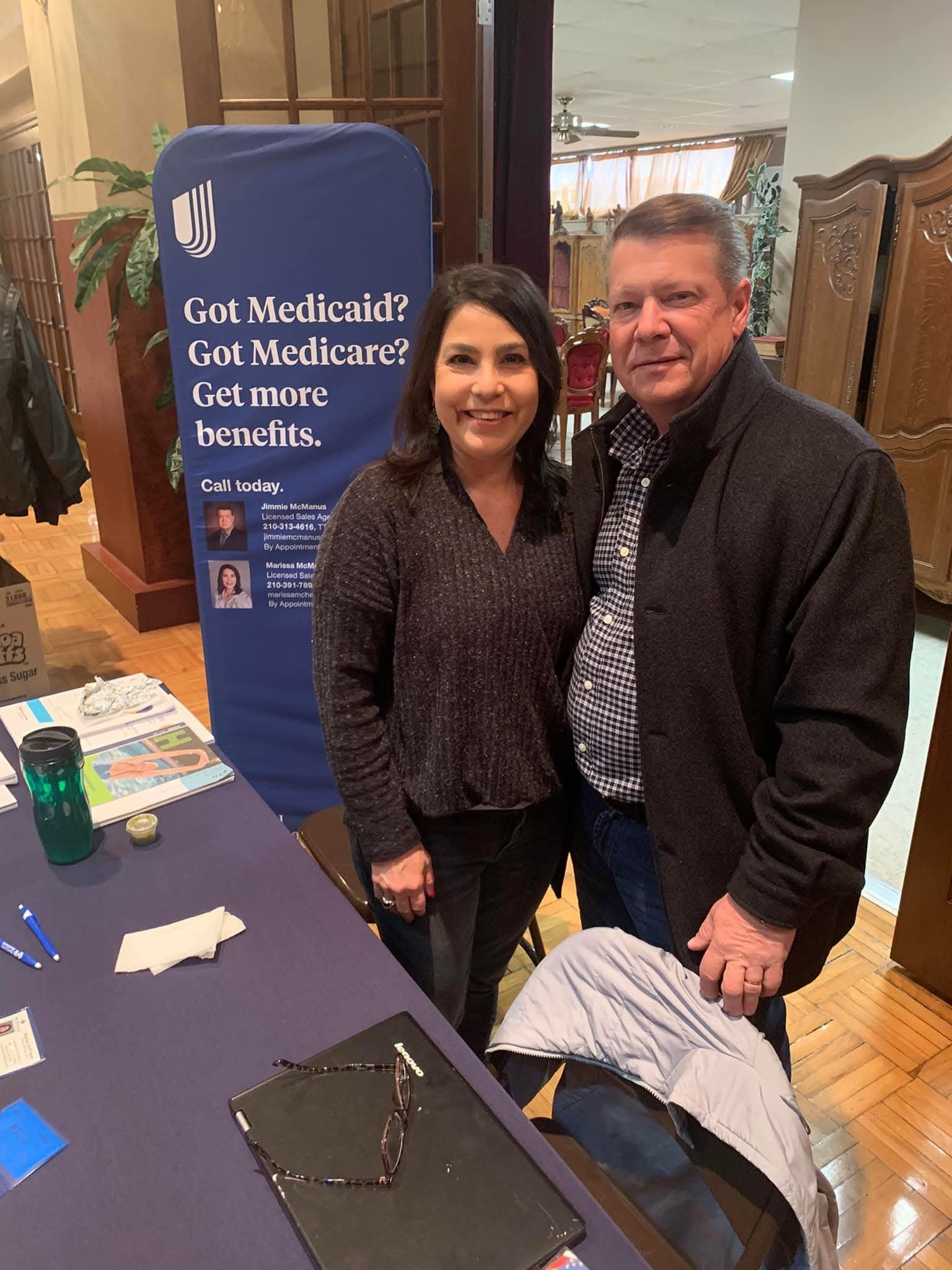 A woman and a man standing together indoors by a table with various items, next to a blue banner that reads 'Got Medicaid? Got Medicare? Get more benefits.'