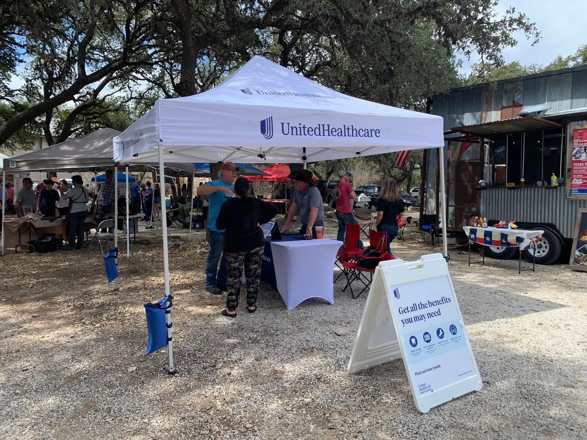 A booth with a white canopy tent bearing the UnitedHealthcare logo is set up outdoors among trees. Several people are interacting and browsing under and around the tent, with a few tables and chairs nearby. The scene appears to be at a community even