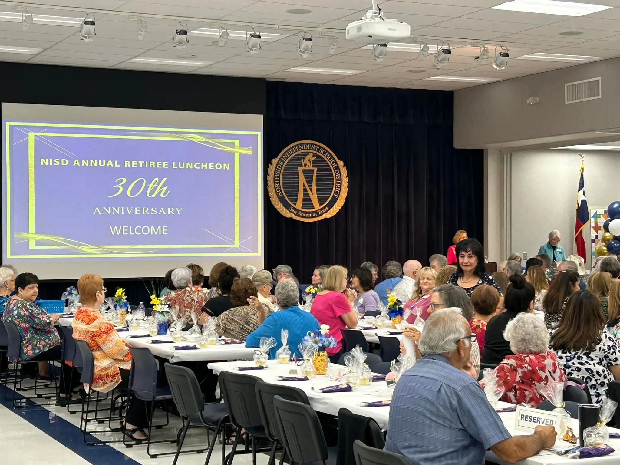 A large room filled with people sitting at decorated tables, attending an annual retired luncheon event. There is a large screen displaying the event details, and a banner with a logo near the stage. Balloons and flowers decorate the tables.
