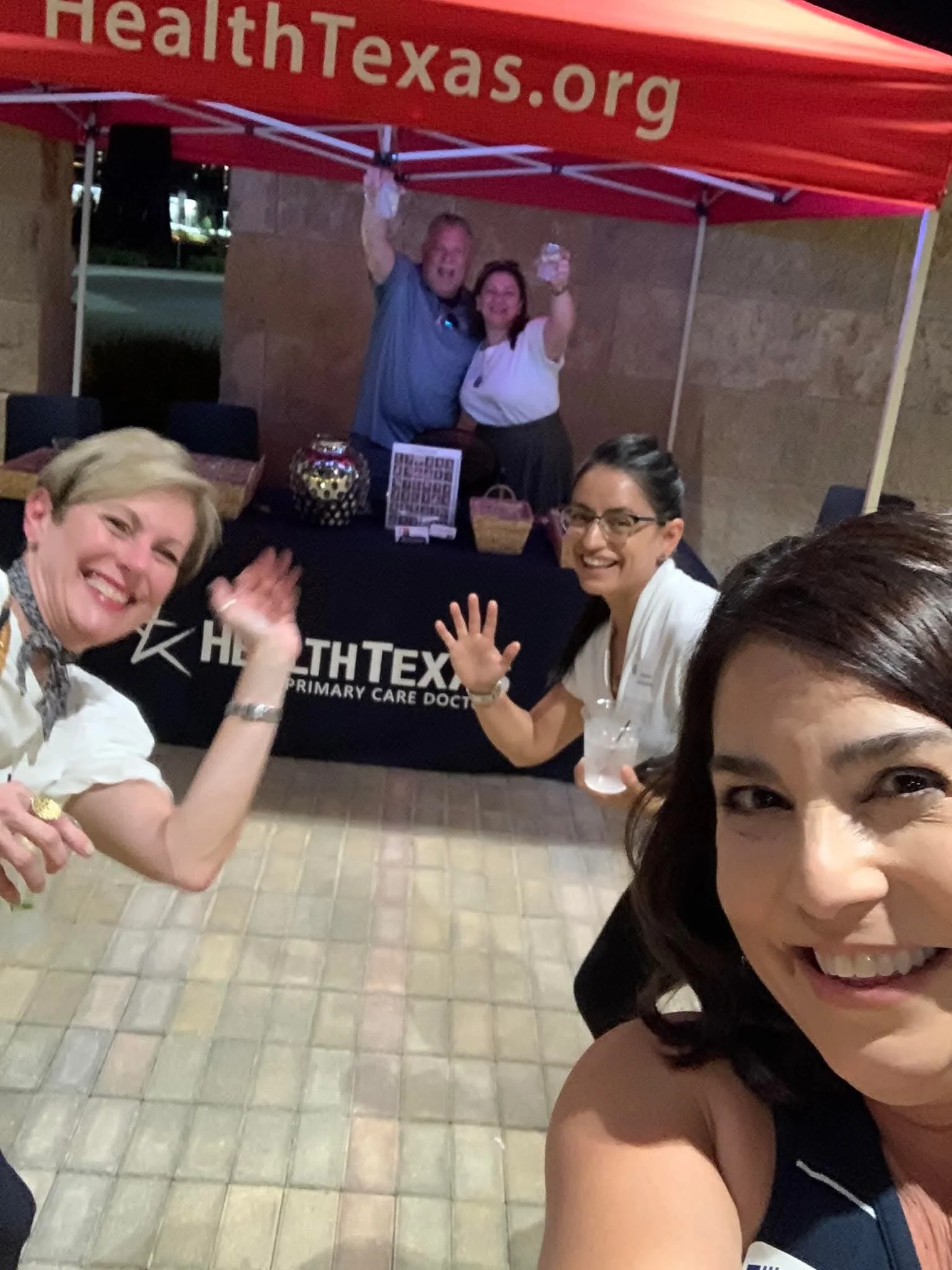 People smiling and waving at a nighttime gathering under a red canopy with 'HealthTexas.org' written on it. Some are holding drinks and wearing name tags.