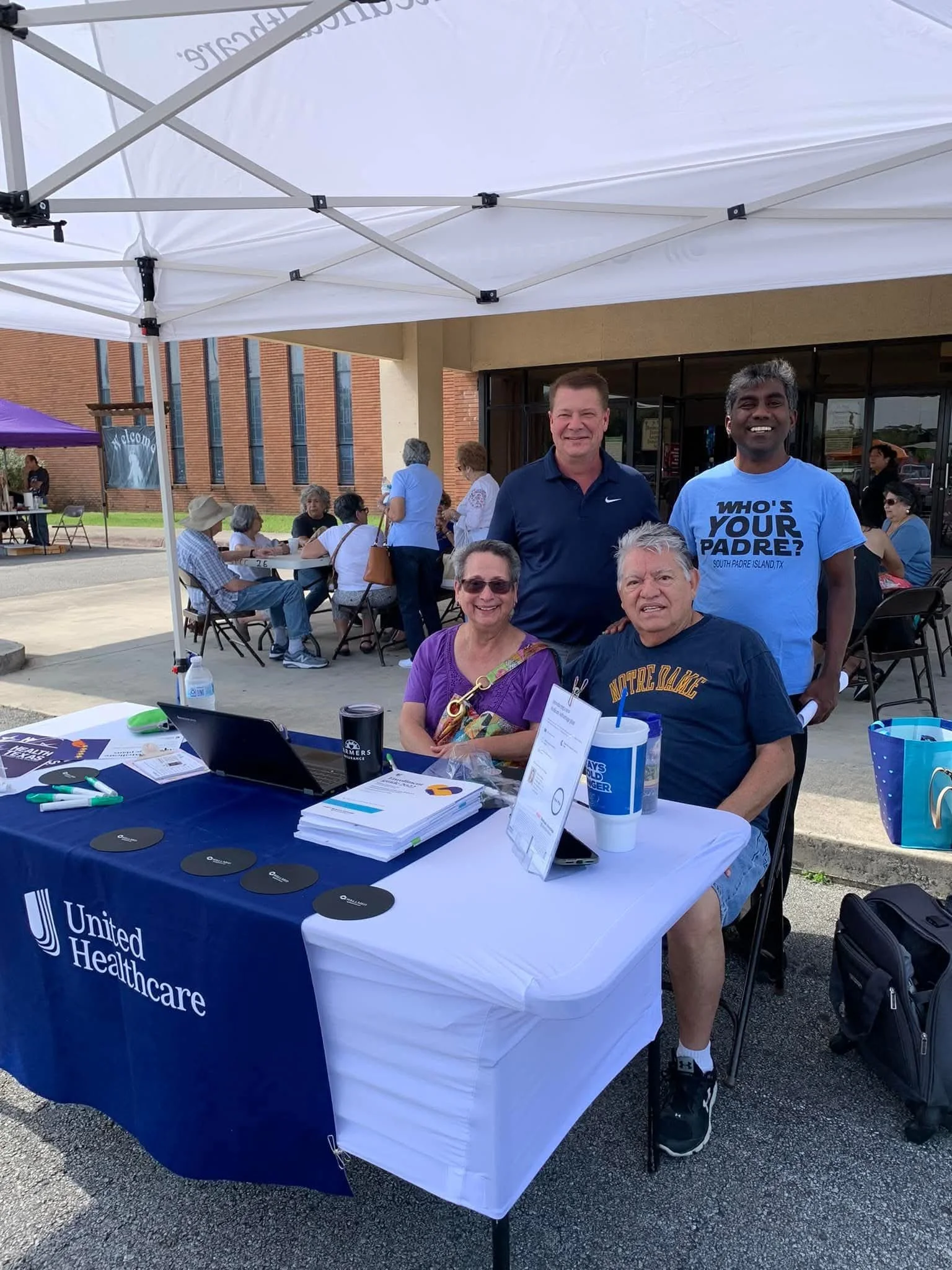 A group of people gathered at an outdoor event, with some seated at tables and others standing. There is a man in a blue Nike shirt, a woman in purple, and a man in a dark blue shirt with the words "North Padre" on it. A table with a United Healthcar