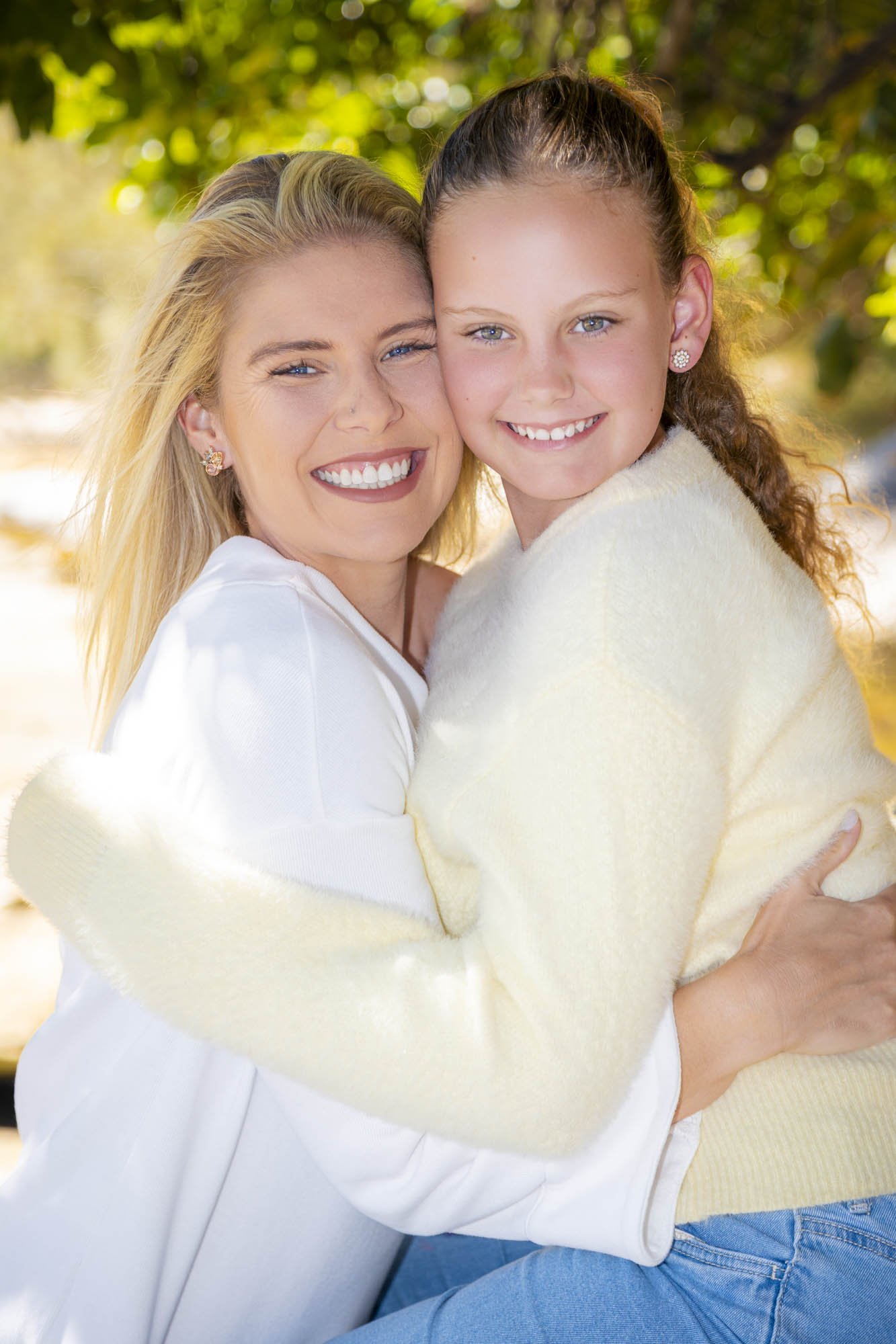 A woman and a girl hugging outdoors, smiling at the camera. The woman has blonde hair and blue eyes, and the girl has light brown hair and blue eyes. They are dressed in light-colored sweaters, with greenery in the background.
