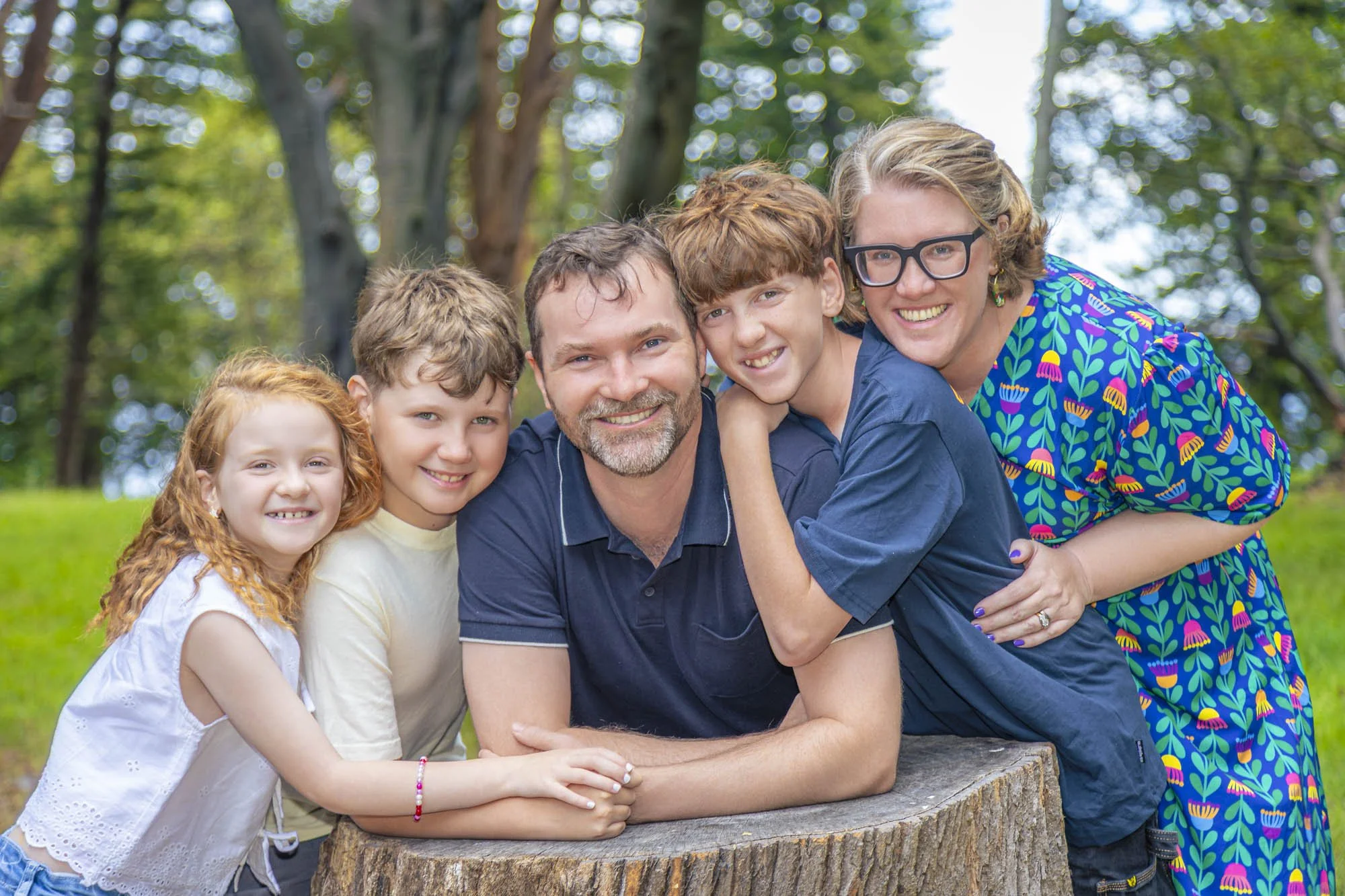 A family of six members wearing casual clothes are outdoors in a park, standing close together around a tree log stump, smiling for the photo.