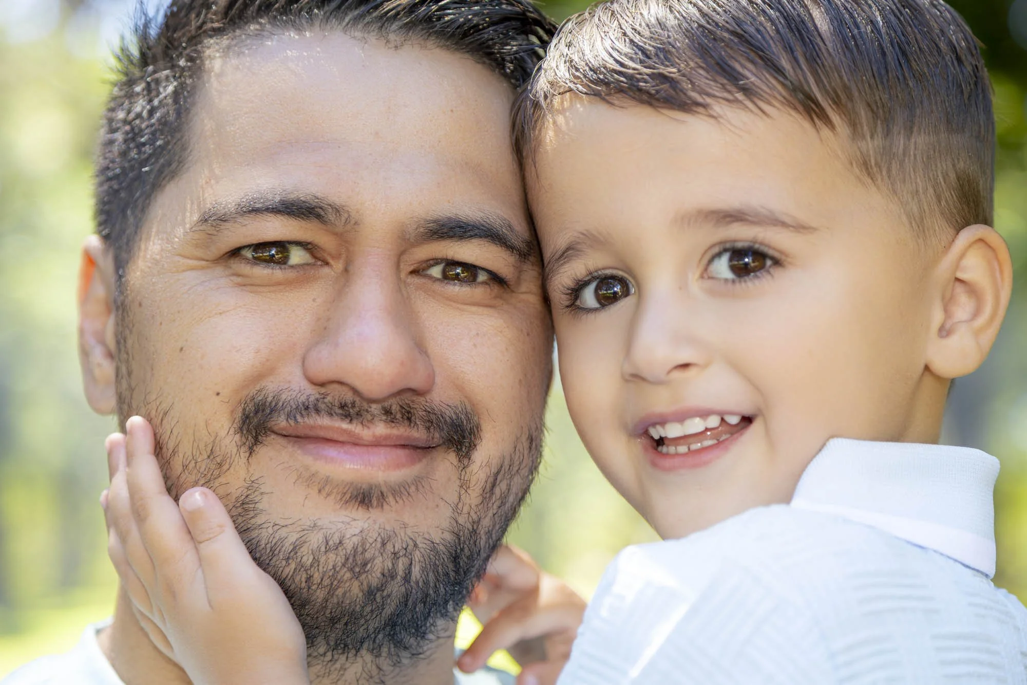 A man and a young boy, both smiling, close-up outdoor portrait. The boy is gently touching the man's face.