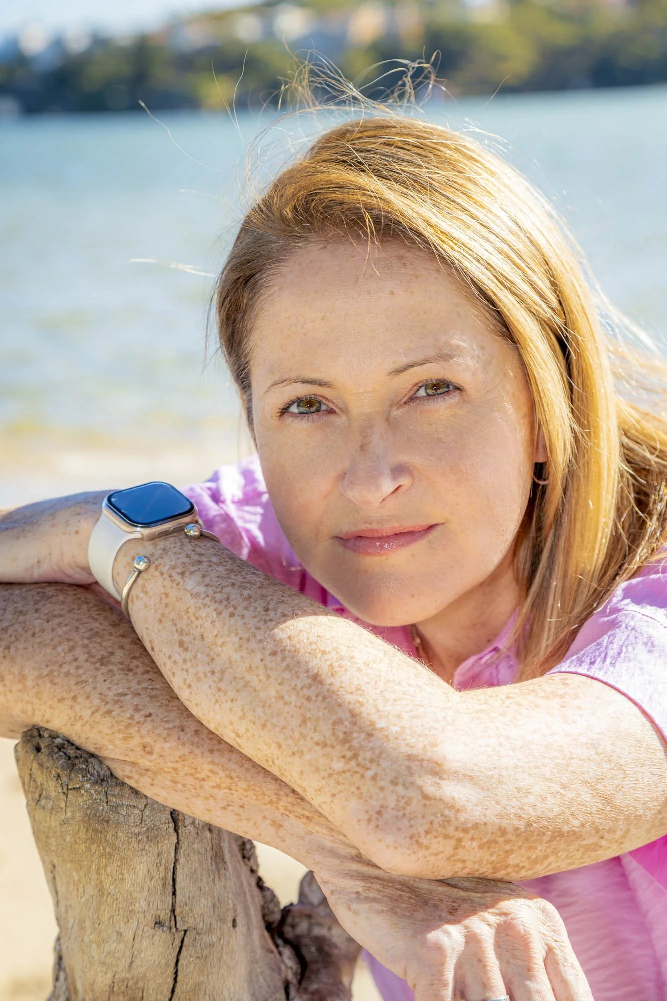 Close-up of a woman with red hair and freckles leaning on a wooden post, looking into the camera with a blue lake and trees in the background.