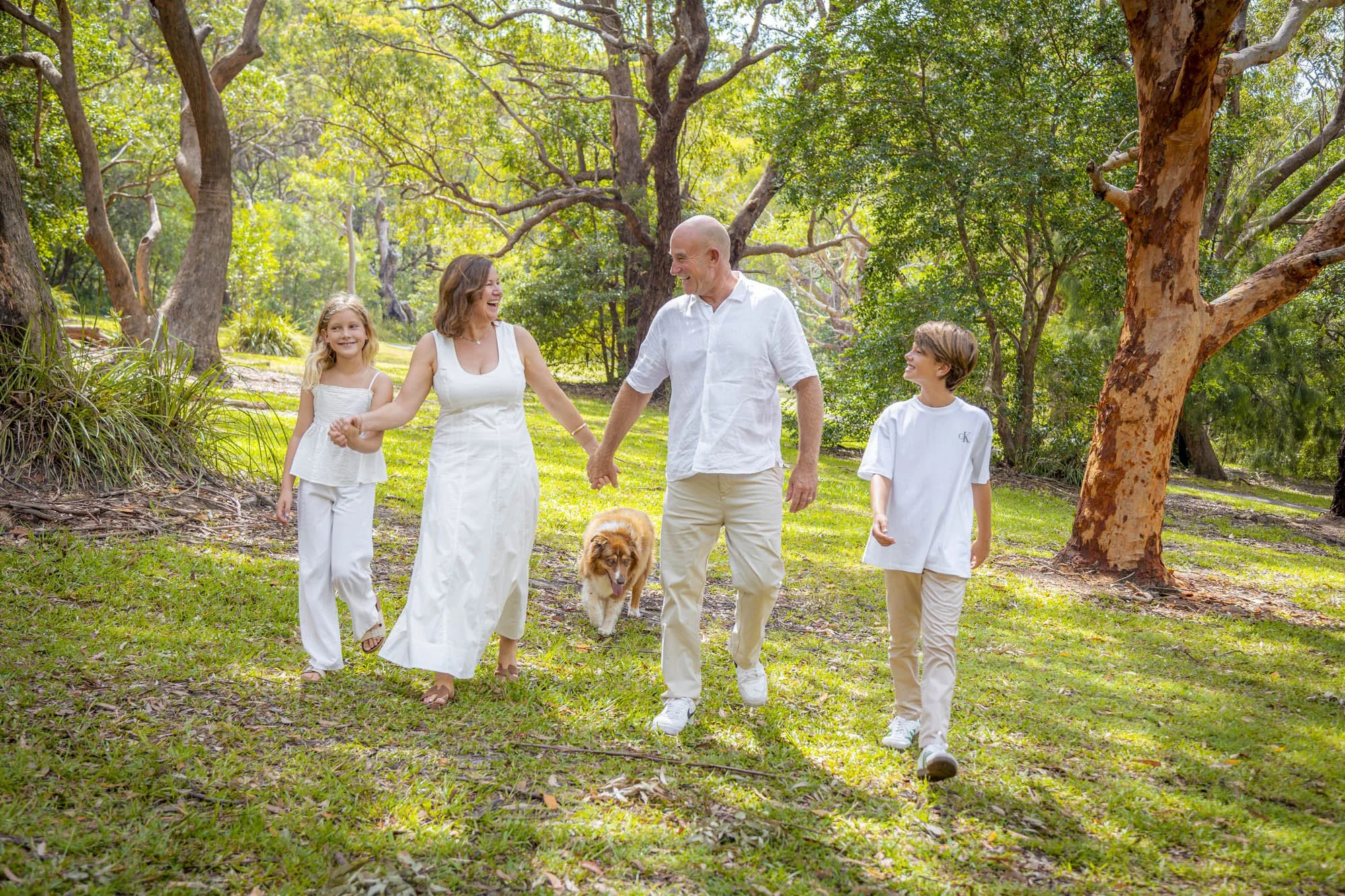 A family walking through a lush green park with tall trees, accompanied by a dog, and enjoying a sunny day with smiles and laughter.
