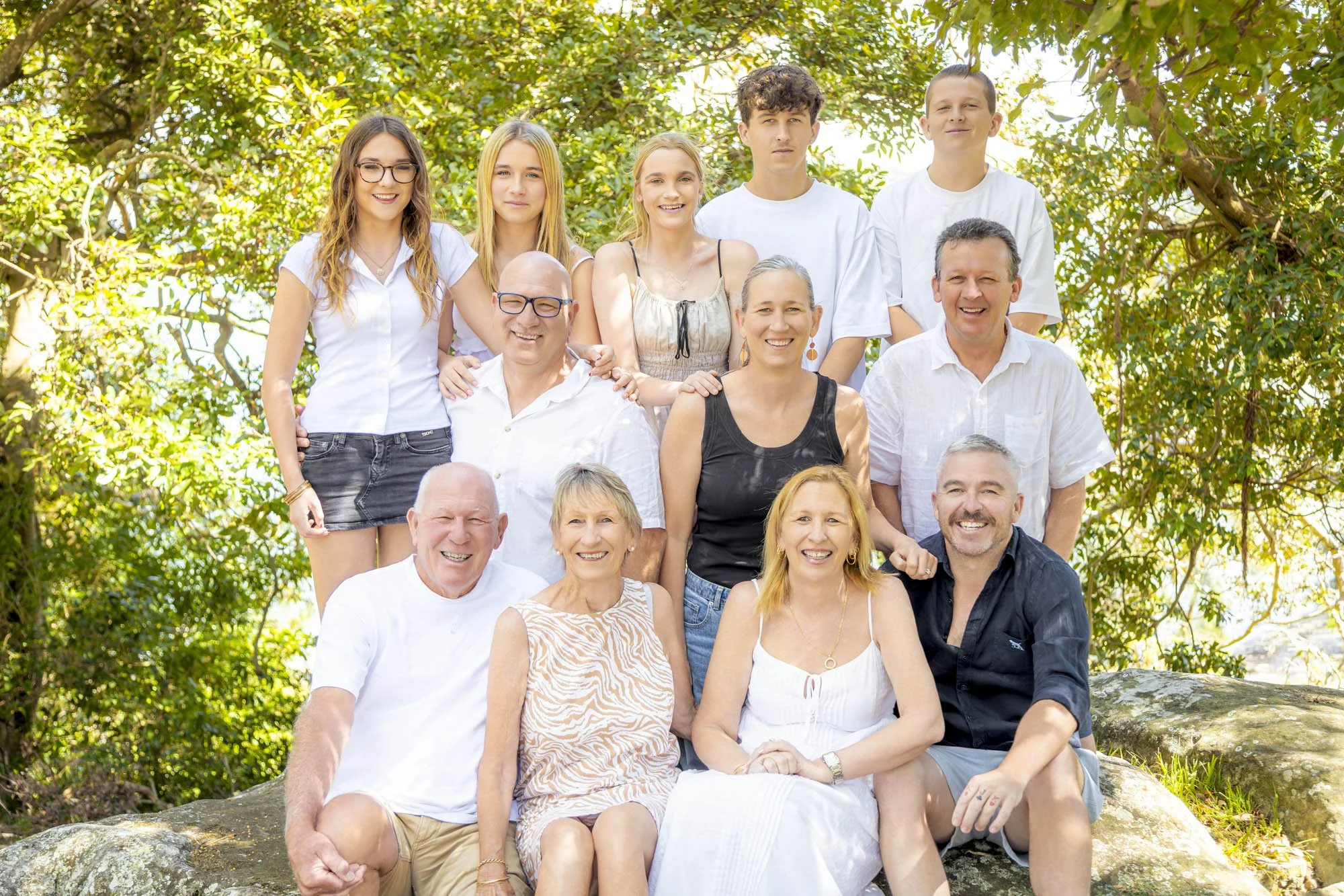 Group of smiling people, including children and older adults, sitting outdoors on rocks with green trees in the background.