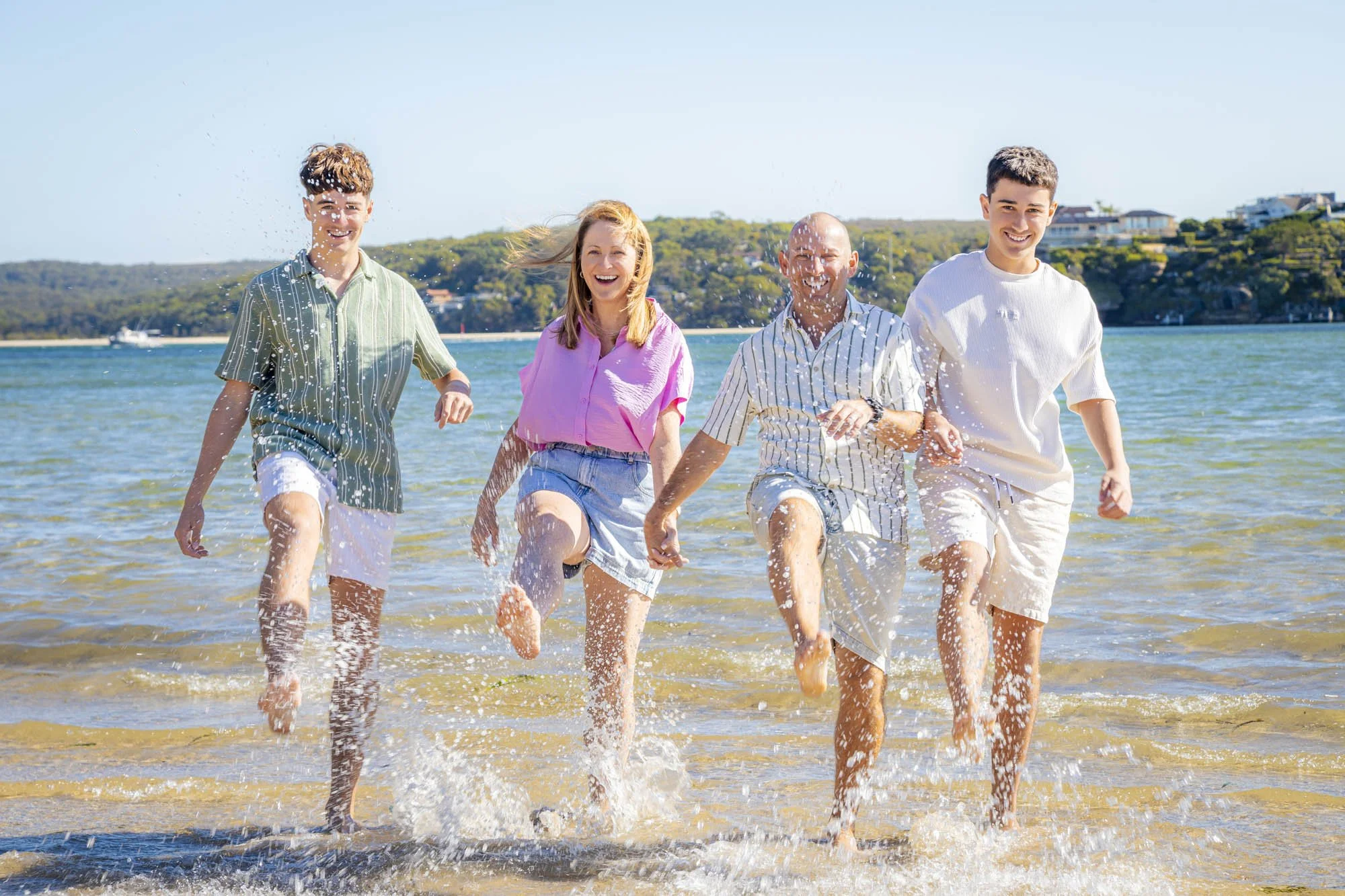 A diverse group of five people, two women and three men, running through shallow water at the beach on a sunny day, smiling and enjoying themselves.