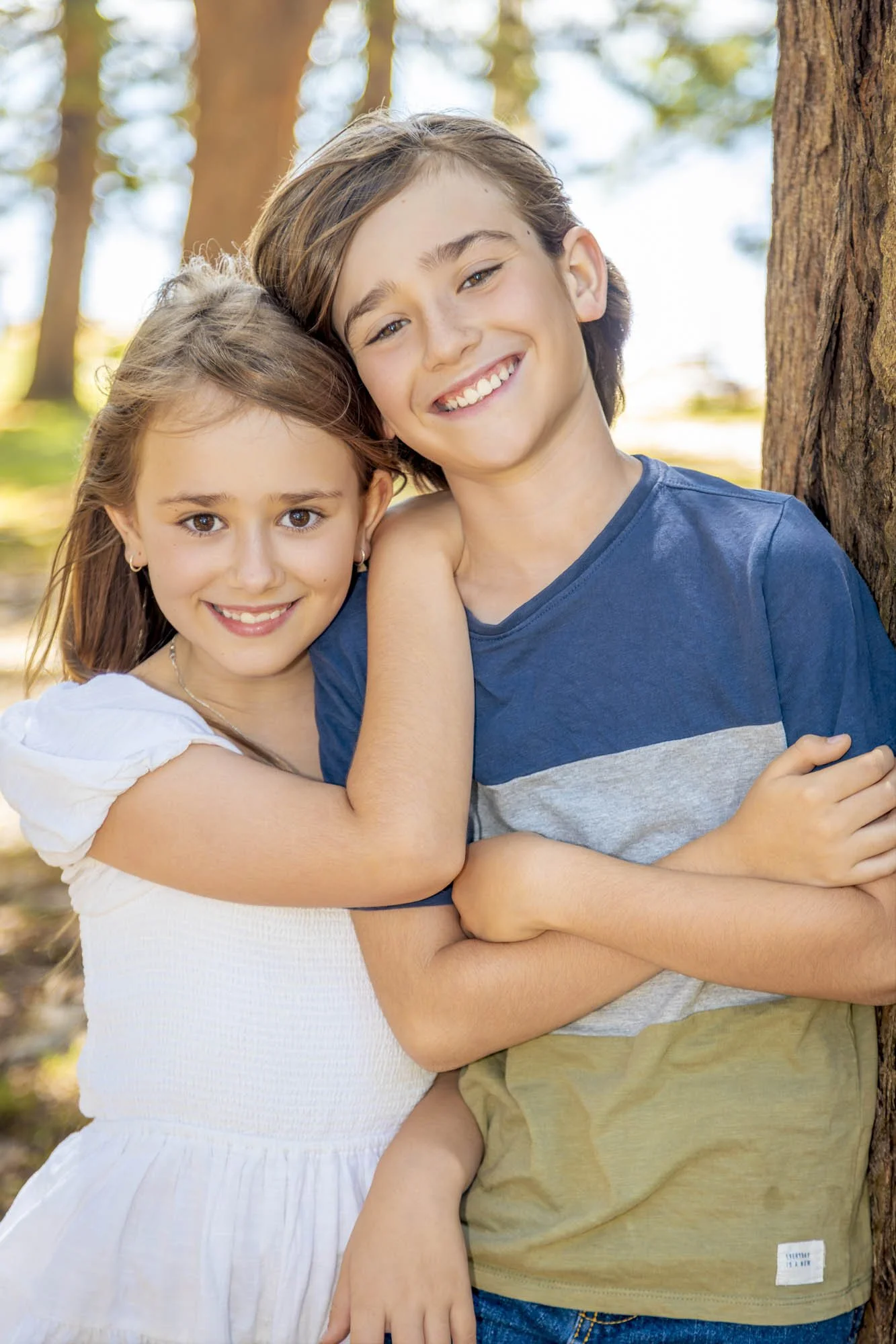 Two kids, a girl and a boy, are smiling and hugging each other outdoors near a tree in a wooded area on a sunny day.
