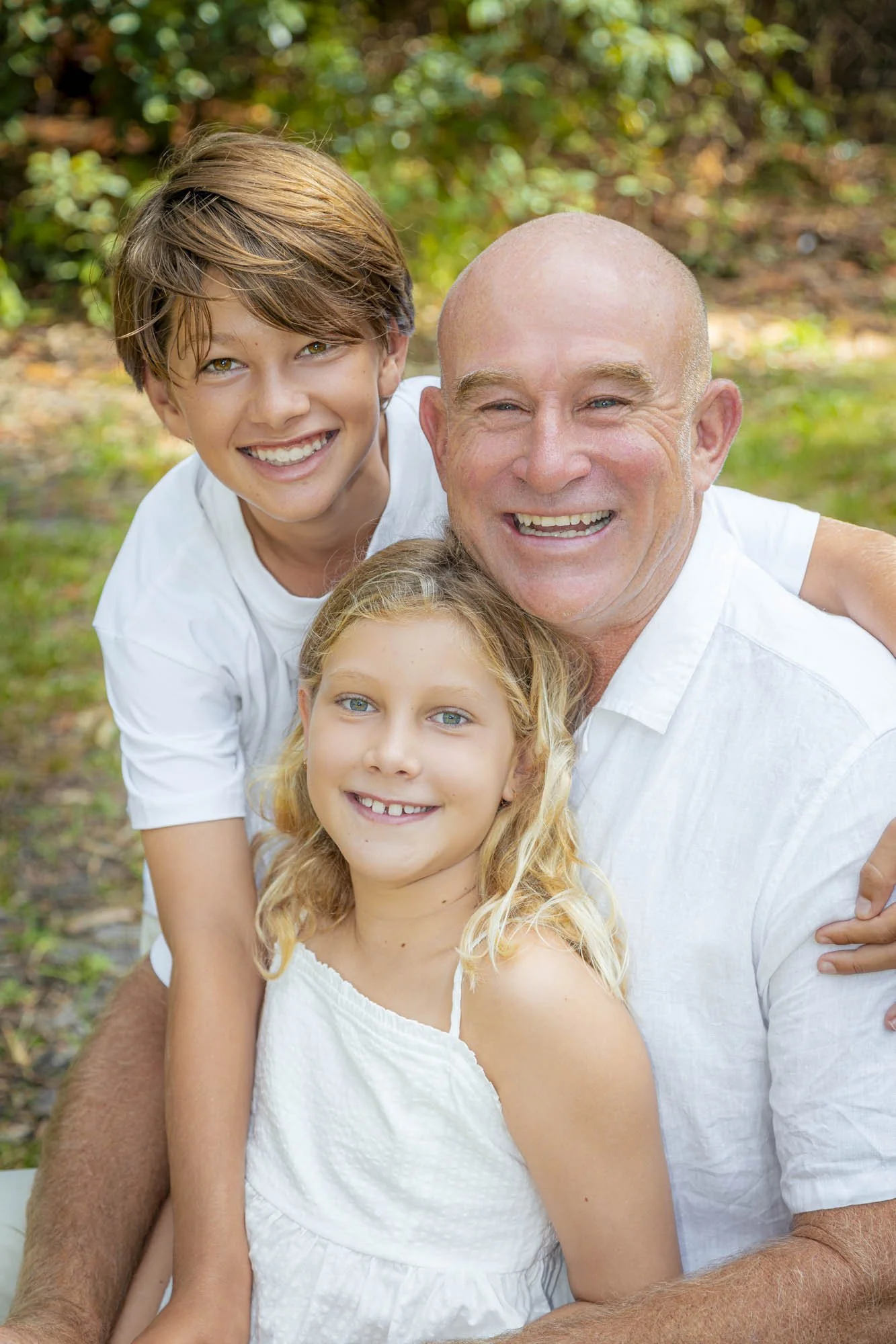 A joyful family outdoors on a sunny day, including a smiling young boy, a happy young girl, and an older man with a bald head, all dressed in white, with a blurred green and brown background.