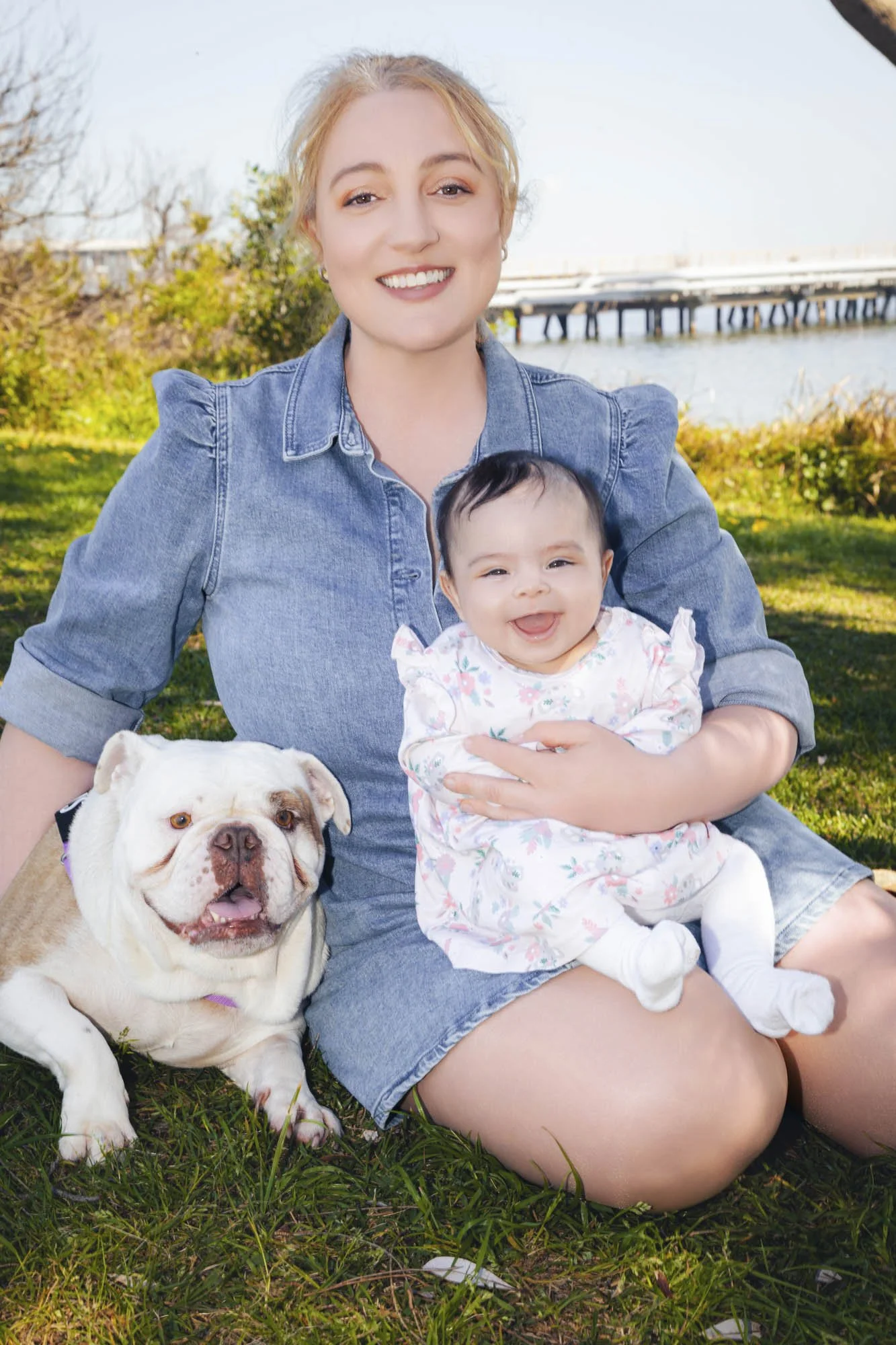 A woman with blonde hair and a baby girl sitting on grass outdoors, smiling at the camera, with a bulldog beside them, a river and a bridge in the background on a sunny day.