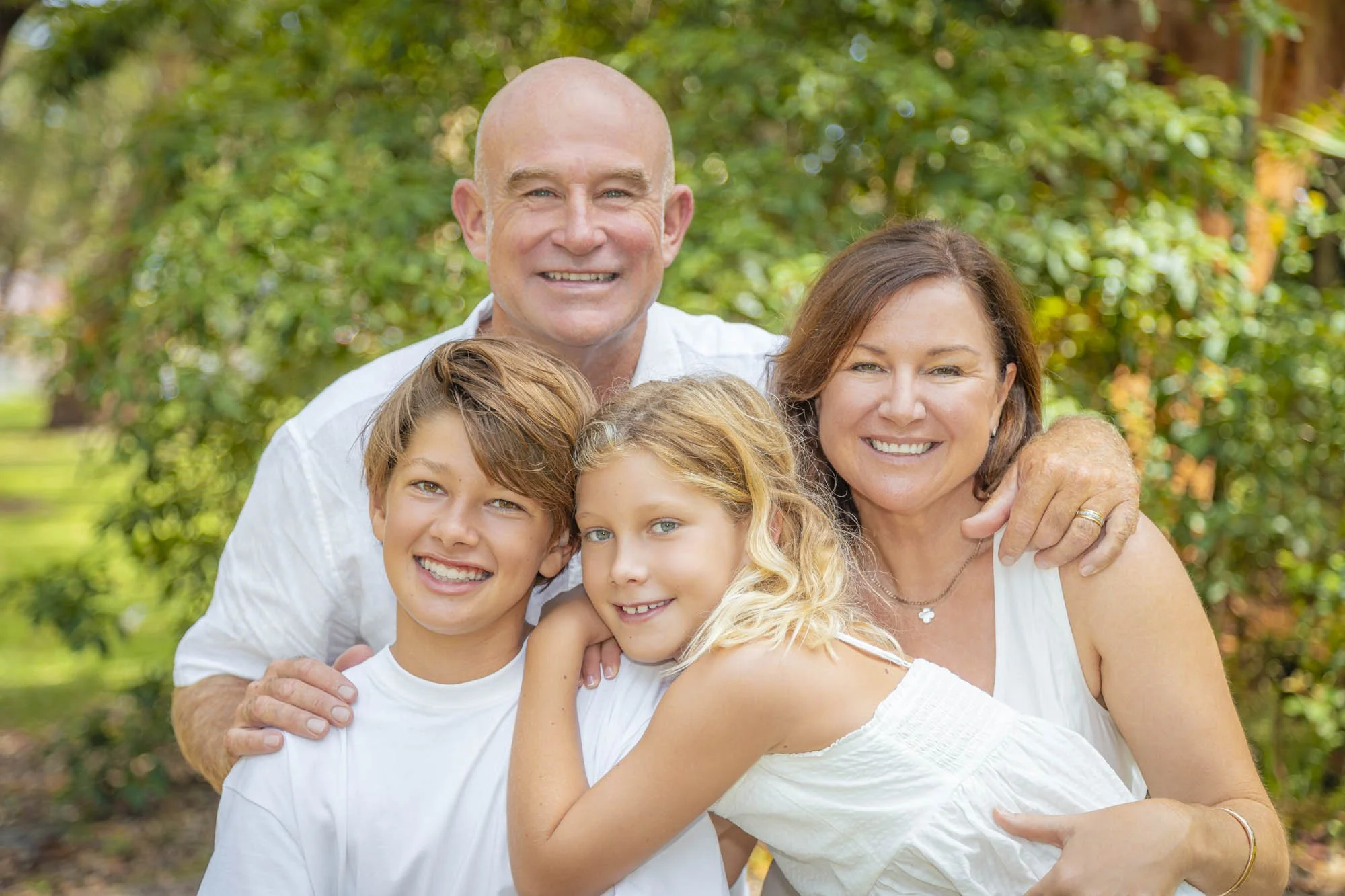 A happy multi-generational family smiling outdoors in a park with green trees in the background. The group includes an older man and woman, and three children, all wearing white shirts.