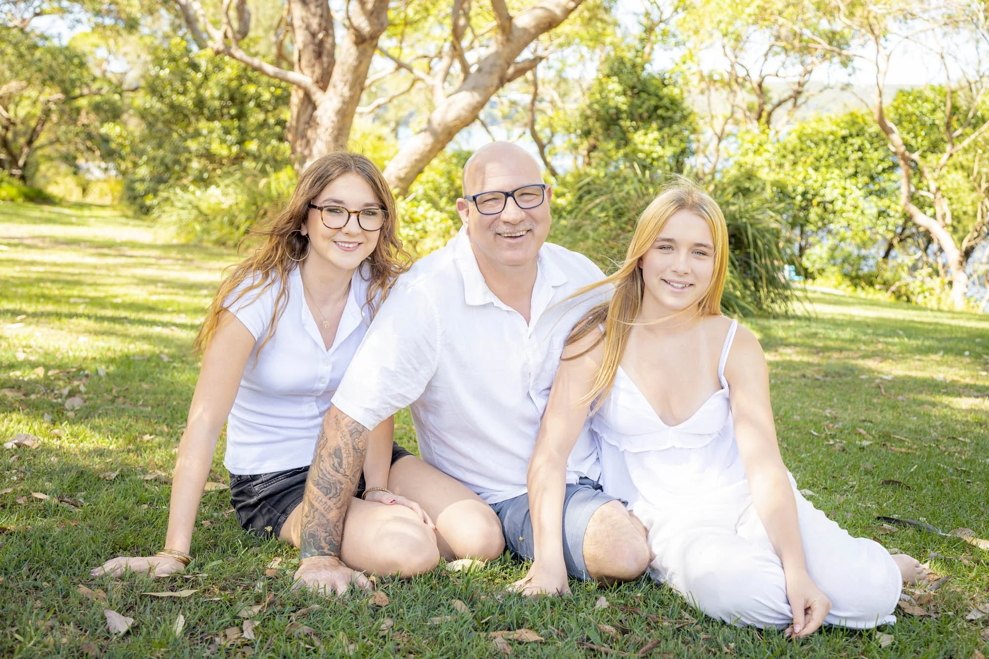 A family of four sitting on the grass in a park with trees in the background. The family includes a young woman with glasses, a bald man with glasses, and a young girl with long blond hair. They are smiling and enjoying a sunny day outdoors.