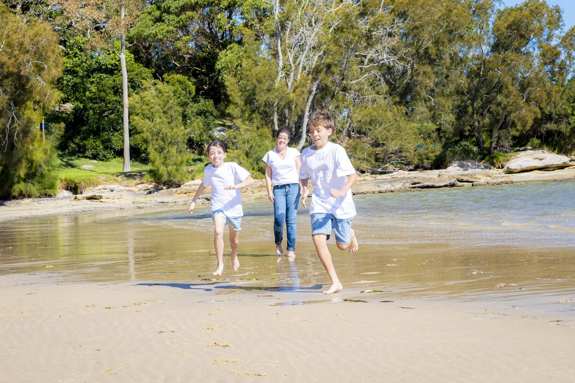 Three children are running on the beach near the water, smiling and enjoying the sunny day with green trees in the background.