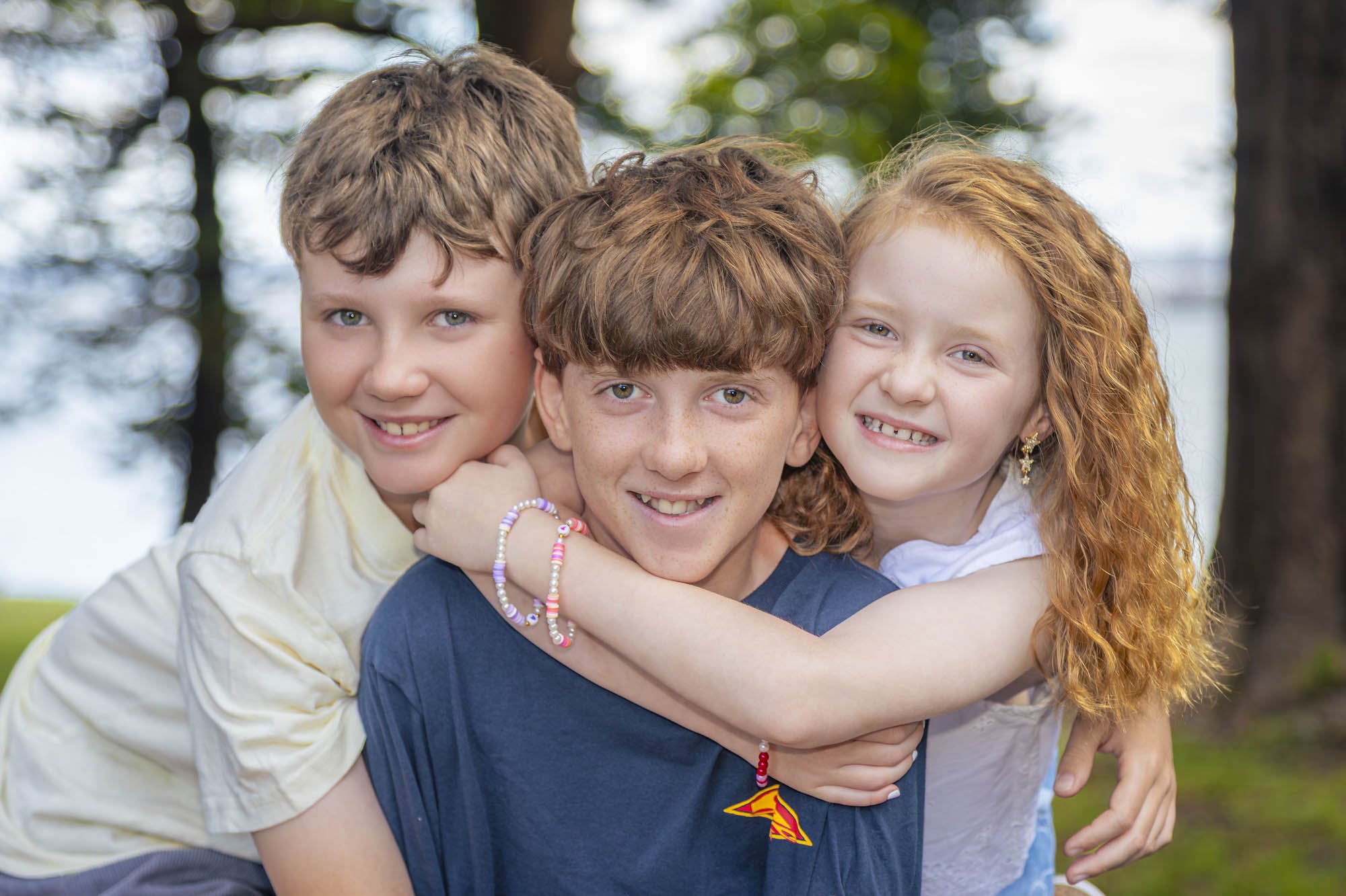 Three children, two girls and one boy, smiling and hugging outdoors in a park with trees and blue sky in the background.