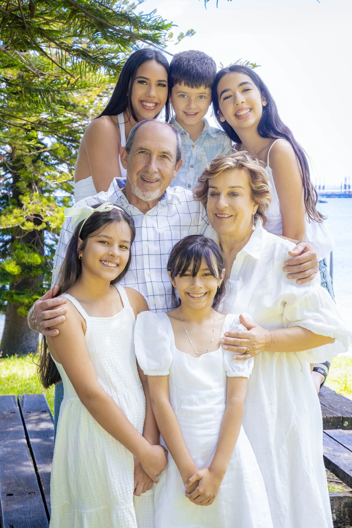 A multi-generational family of nine people smiling outdoors on a sunny day by a body of water, with trees and picnic tables in the background, dressed in white and light-colored clothing.