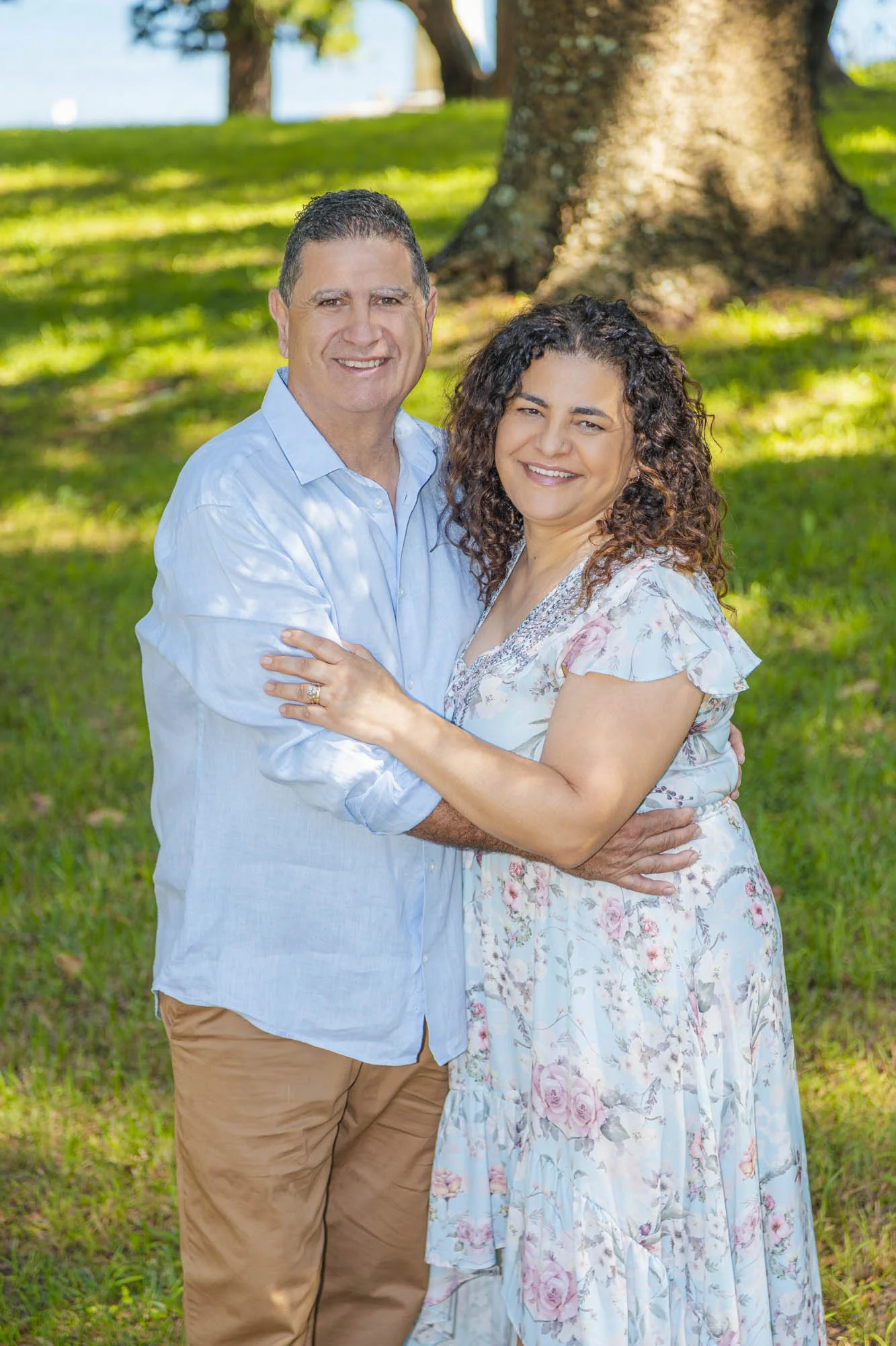 A smiling middle-aged man and woman hugging outdoors in a park with green grass and trees.
