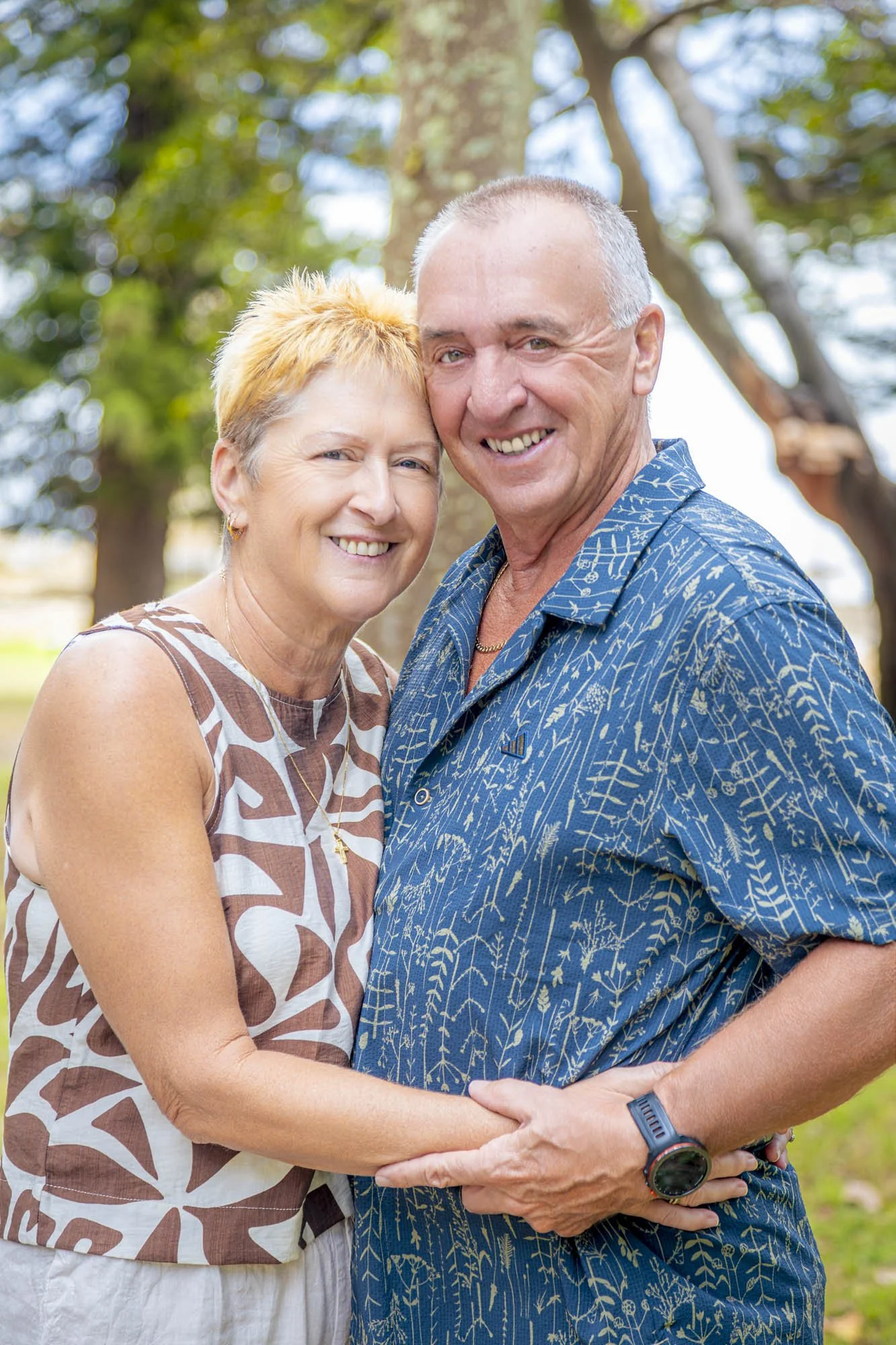 A smiling older couple holding hands outdoors with trees in the background.
