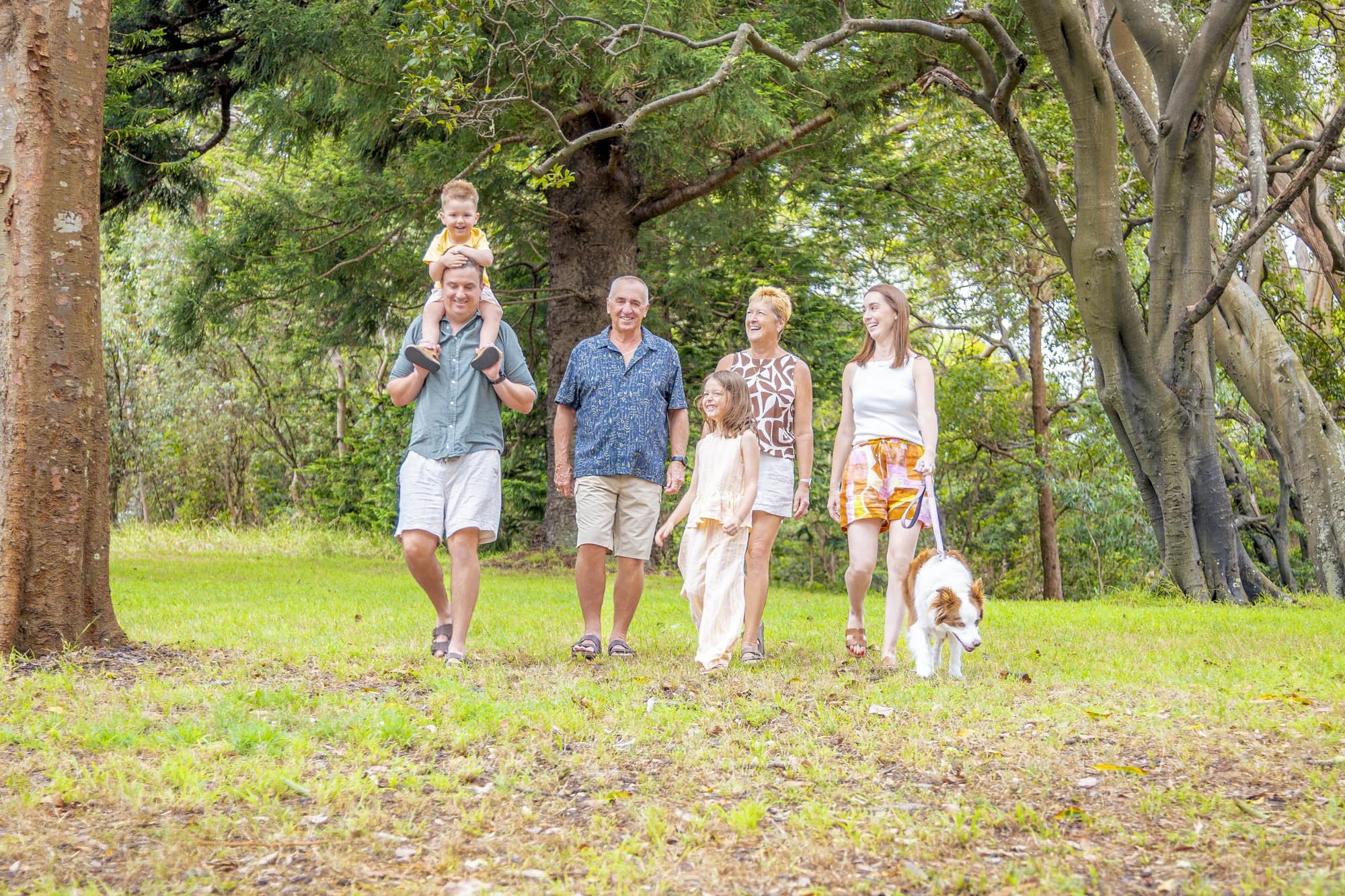 Family walking on a grassy trail in a lush park, with a dog leading, during daytime.