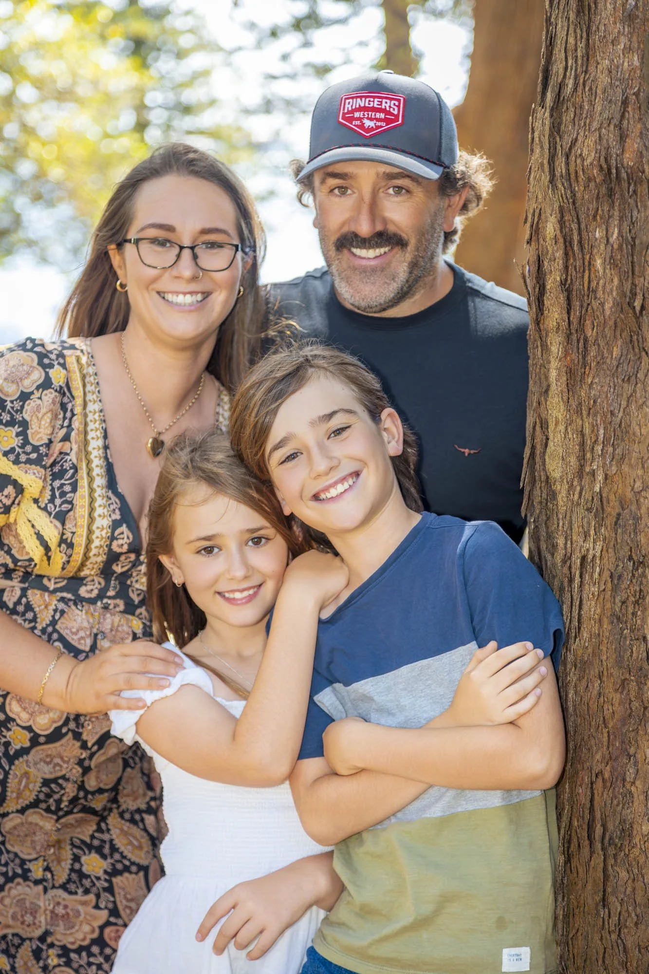A smiling family of five, including two young girls and two adults, poses outdoors next to a tree during daytime. The woman has glasses and is wearing a patterned dress, while the man is wearing a baseball cap and a black t-shirt. The children are ha