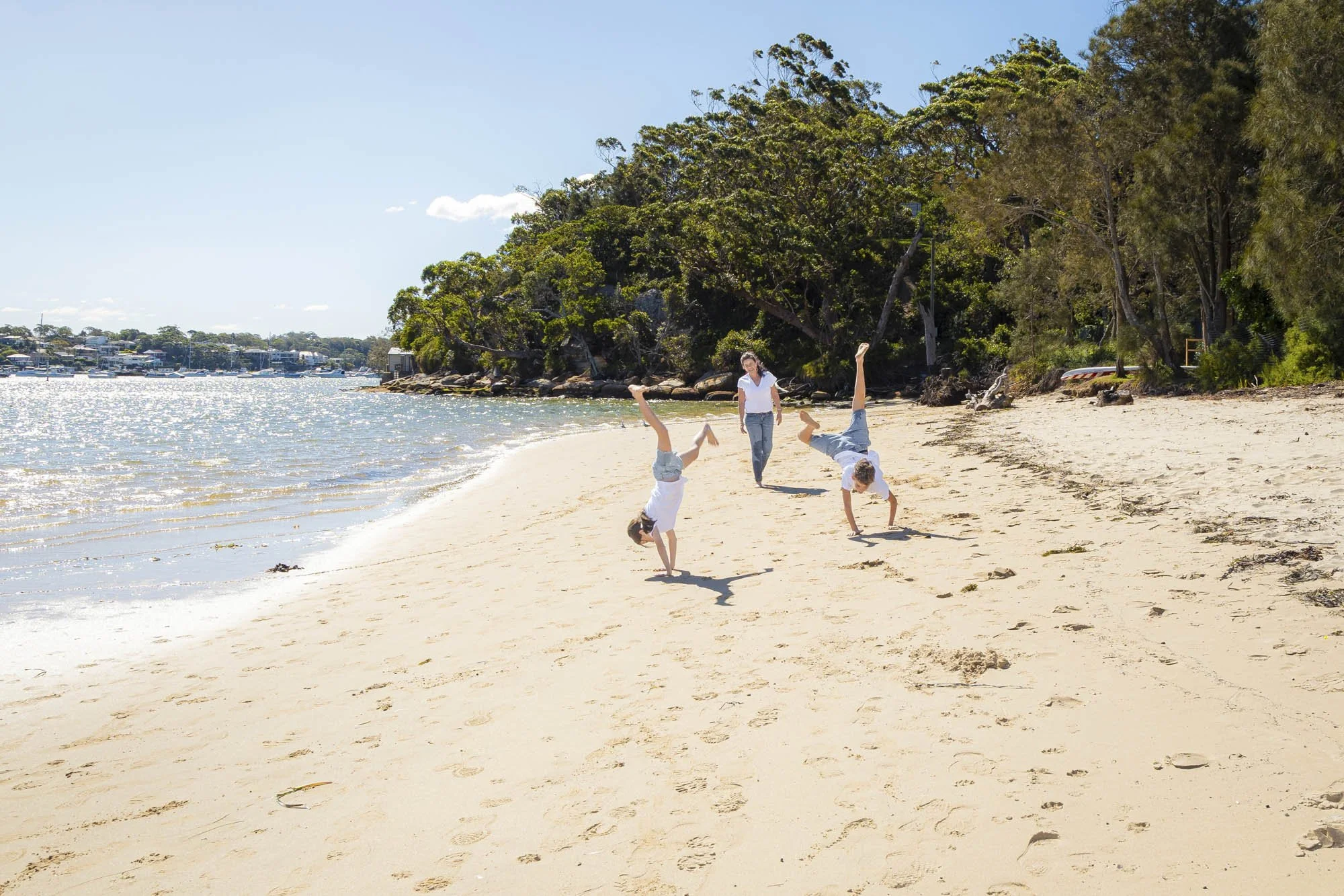 A woman and three children playing and doing cartwheels on a sandy beach near the water, with green trees and boats in the background on a sunny day.