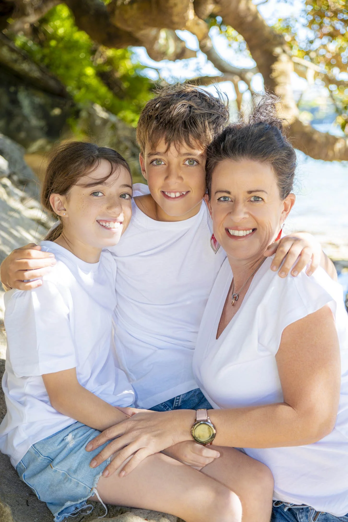 A woman and two children sitting close together outdoors during daytime, smiling at the camera, with trees and bright sky in the background.