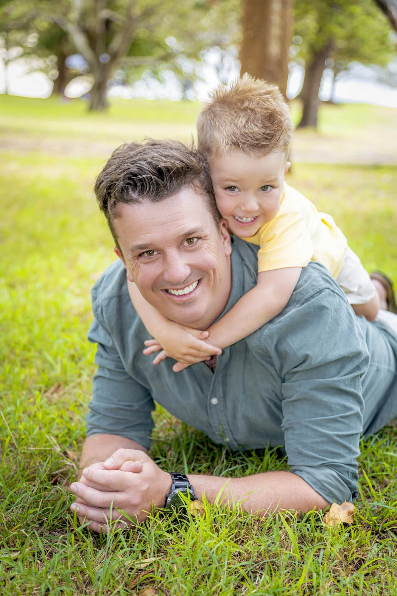 A man with short dark hair and wearing a blue shirt is laying on the grass outdoors, supporting his head with his hands and smiling at the camera. A young boy with red hair and a yellow shirt is riding on his back, also smiling. The background featur