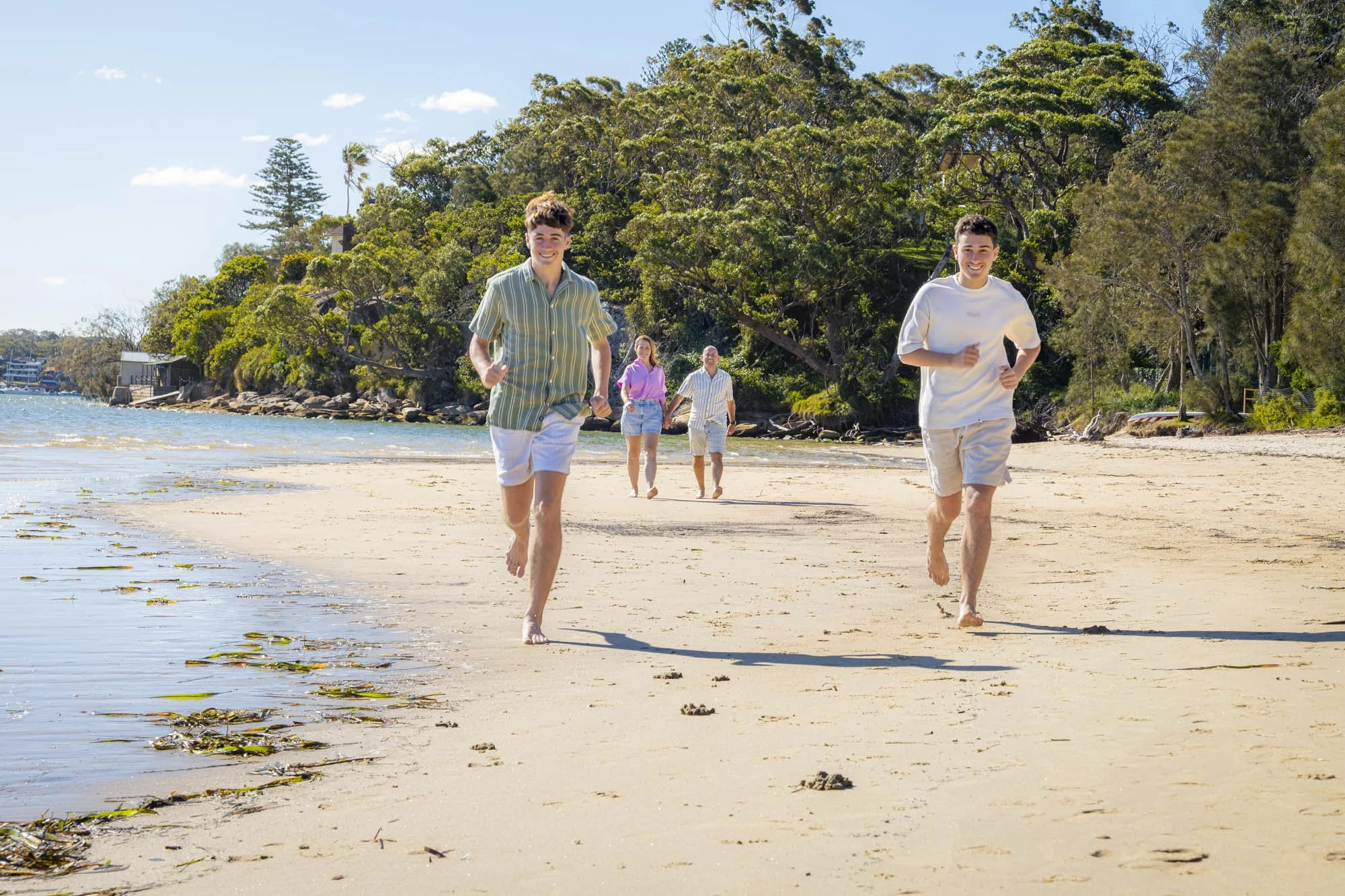 Two young men are running on a sandy beach, smiling and barefoot. Behind them, an older couple walks along the shoreline. The beach is bordered by lush green trees and the sky is clear with a few clouds.