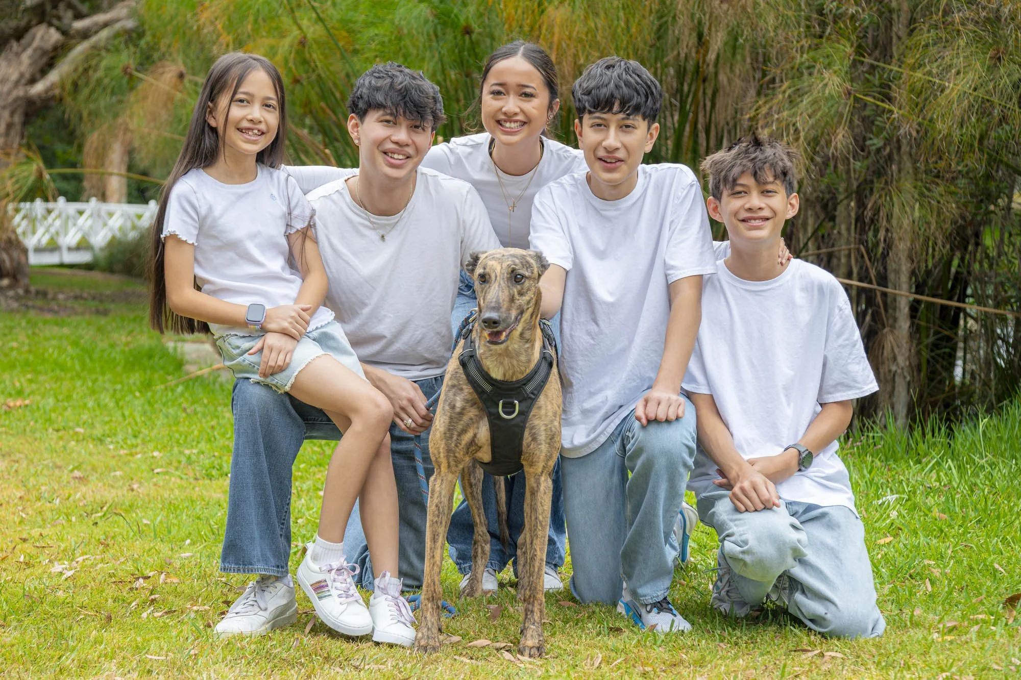 A group of five children and a dog outdoors in a grassy area with trees in the background. The children are smiling and wearing white shirts.