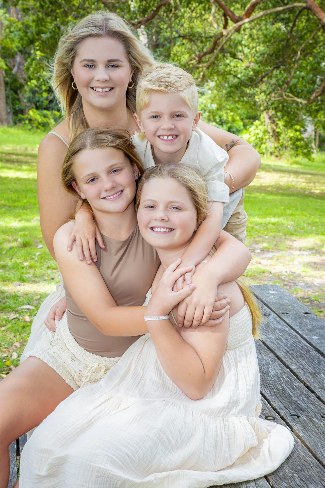 A woman and four children sitting and hugging outdoors on a wooden bench surrounded by trees and green grass.
