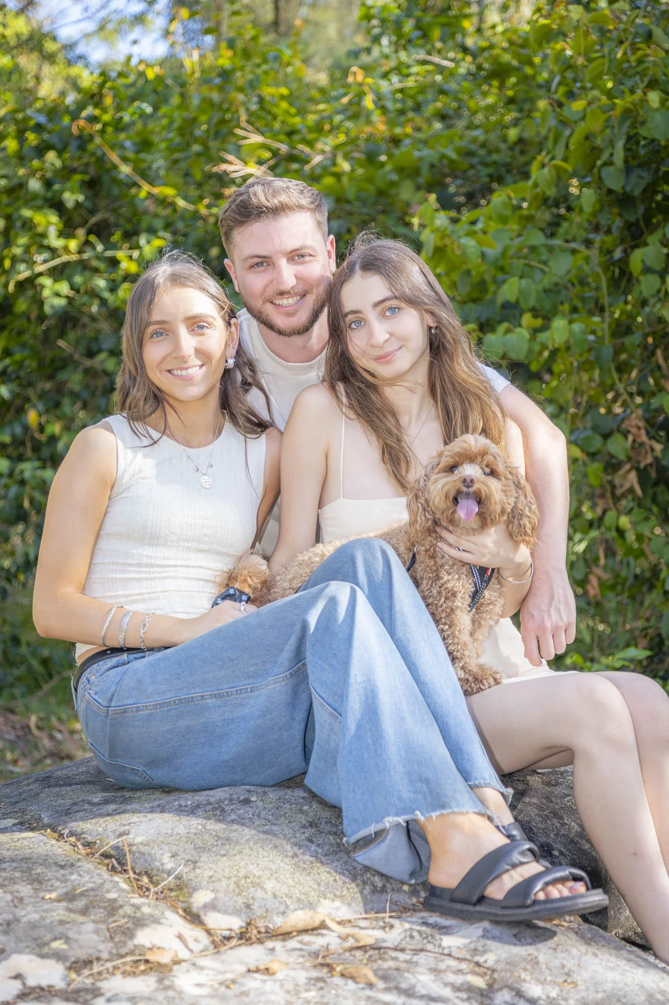 A group of three young adults and a small brown poodle sitting outdoors on a large rock, surrounded by green trees and foliage, all smiling at the camera.