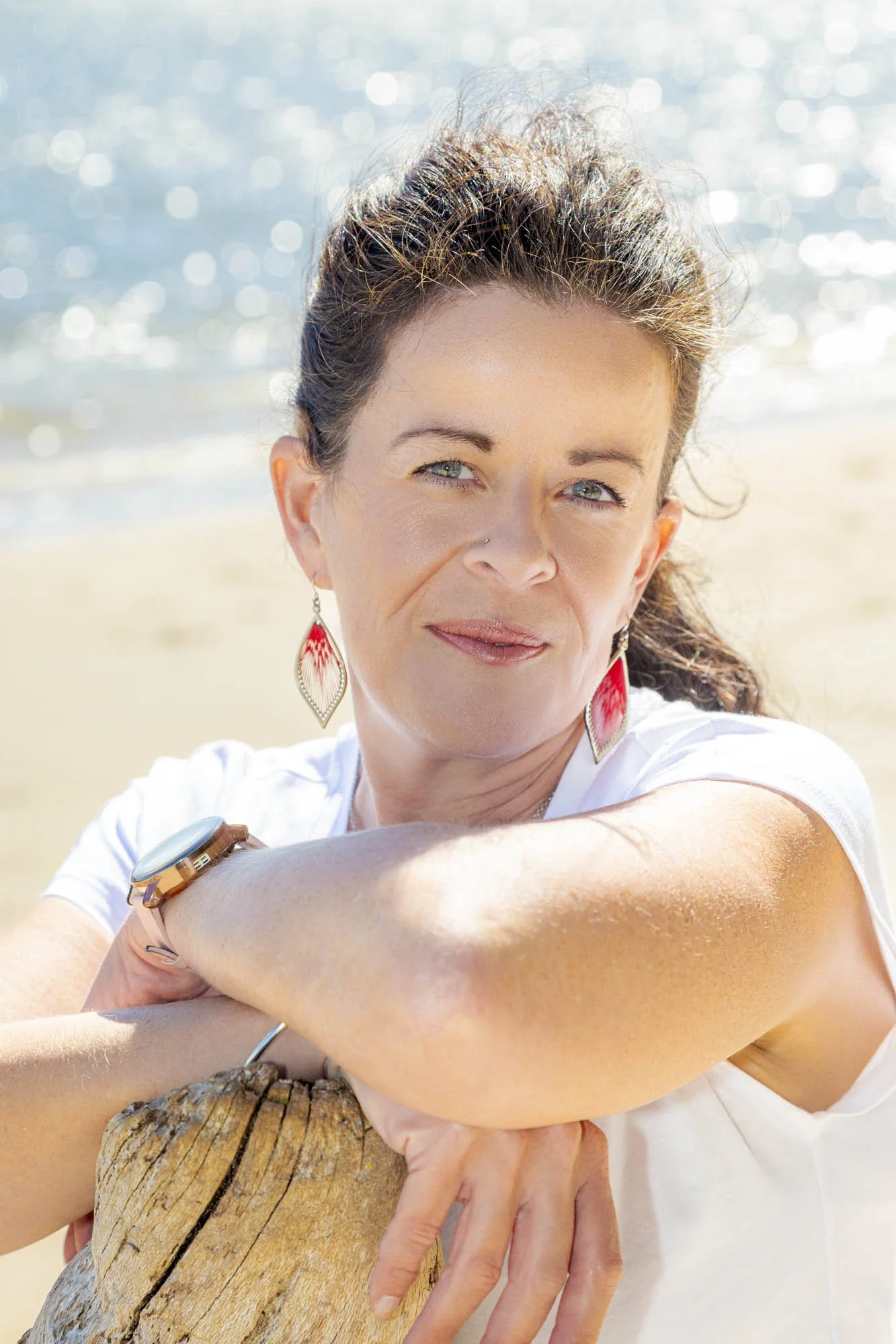 A woman with blue eyes and brown hair, wearing earrings and a white shirt, is hugging a large tree trunk on a sunny beach with the ocean in the background.