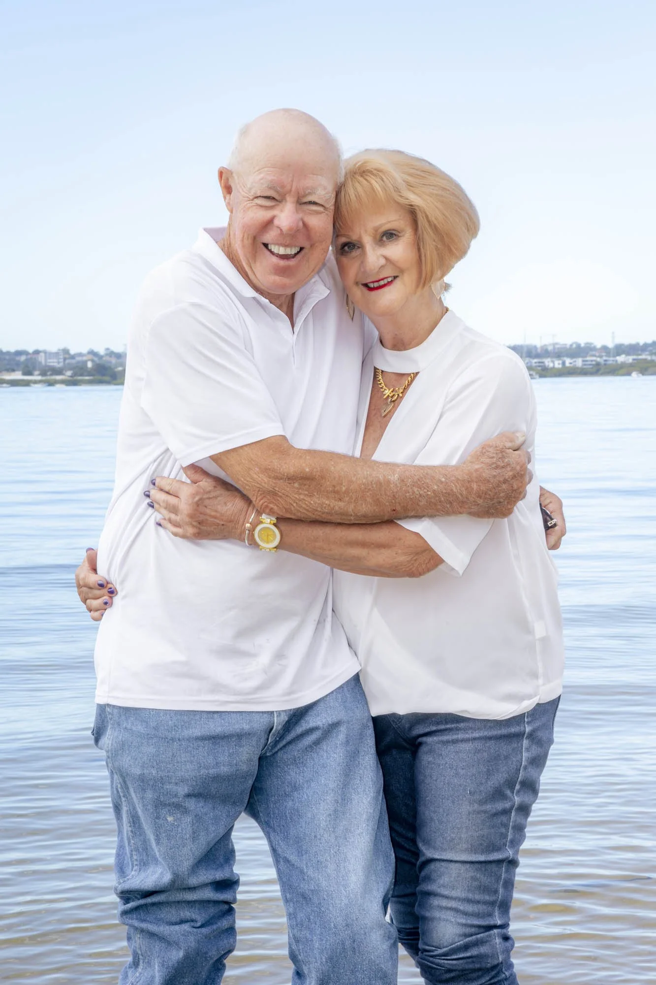 A happy older couple hugging by the water, both wearing white tops and jeans, with a city skyline in the background.