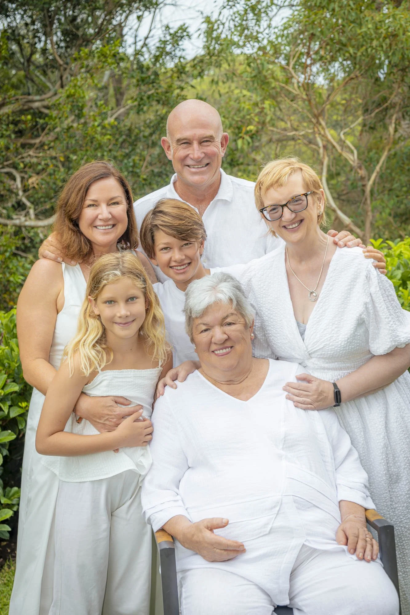 A multigenerational family group portrait outdoors, with eight people including children, middle-aged, and elderly, all dressed in white, smiling in front of green trees.