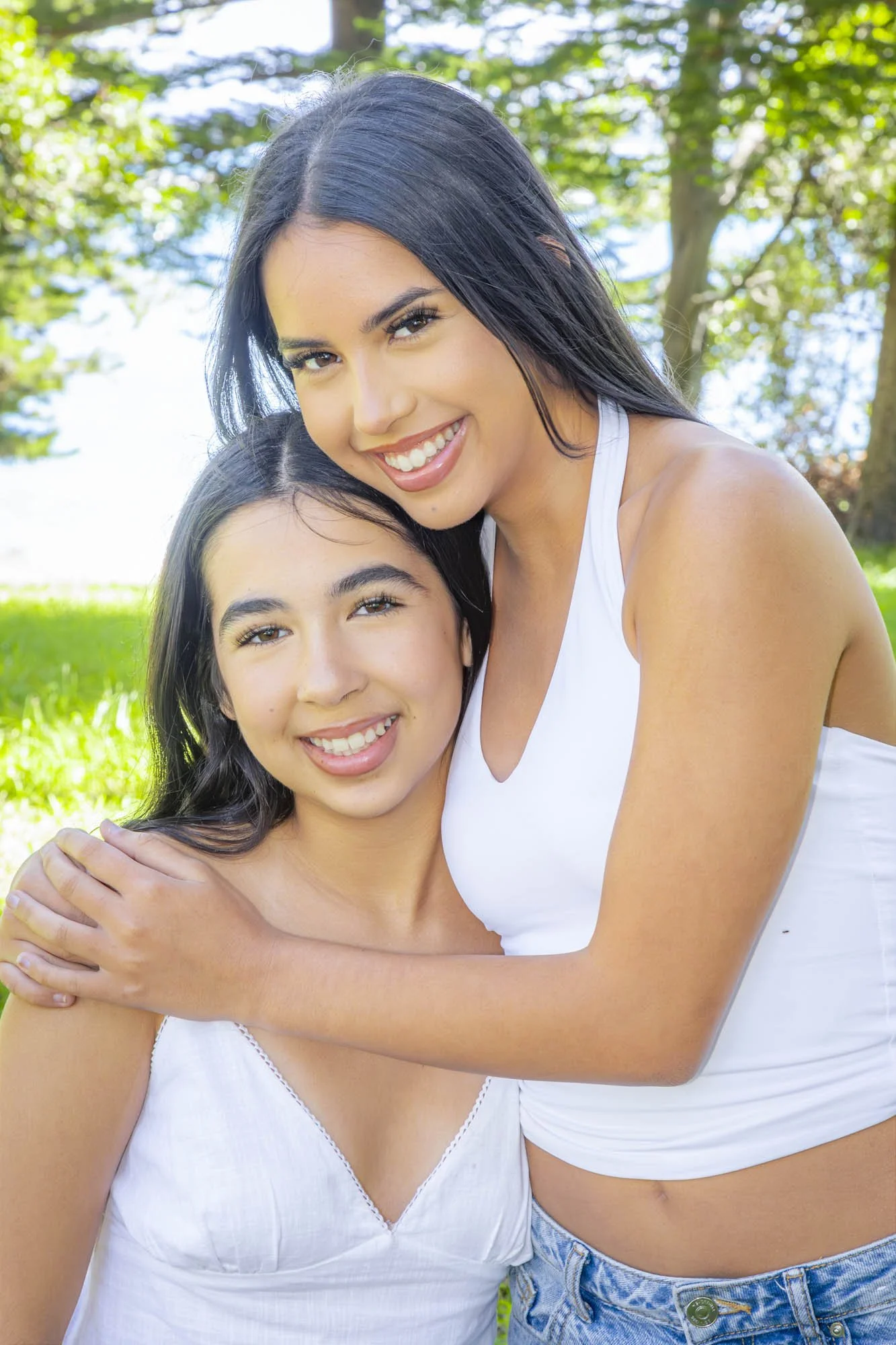 A woman and a girl hugging outdoors in a park, both smiling at the camera, with trees and green grass in the background.
