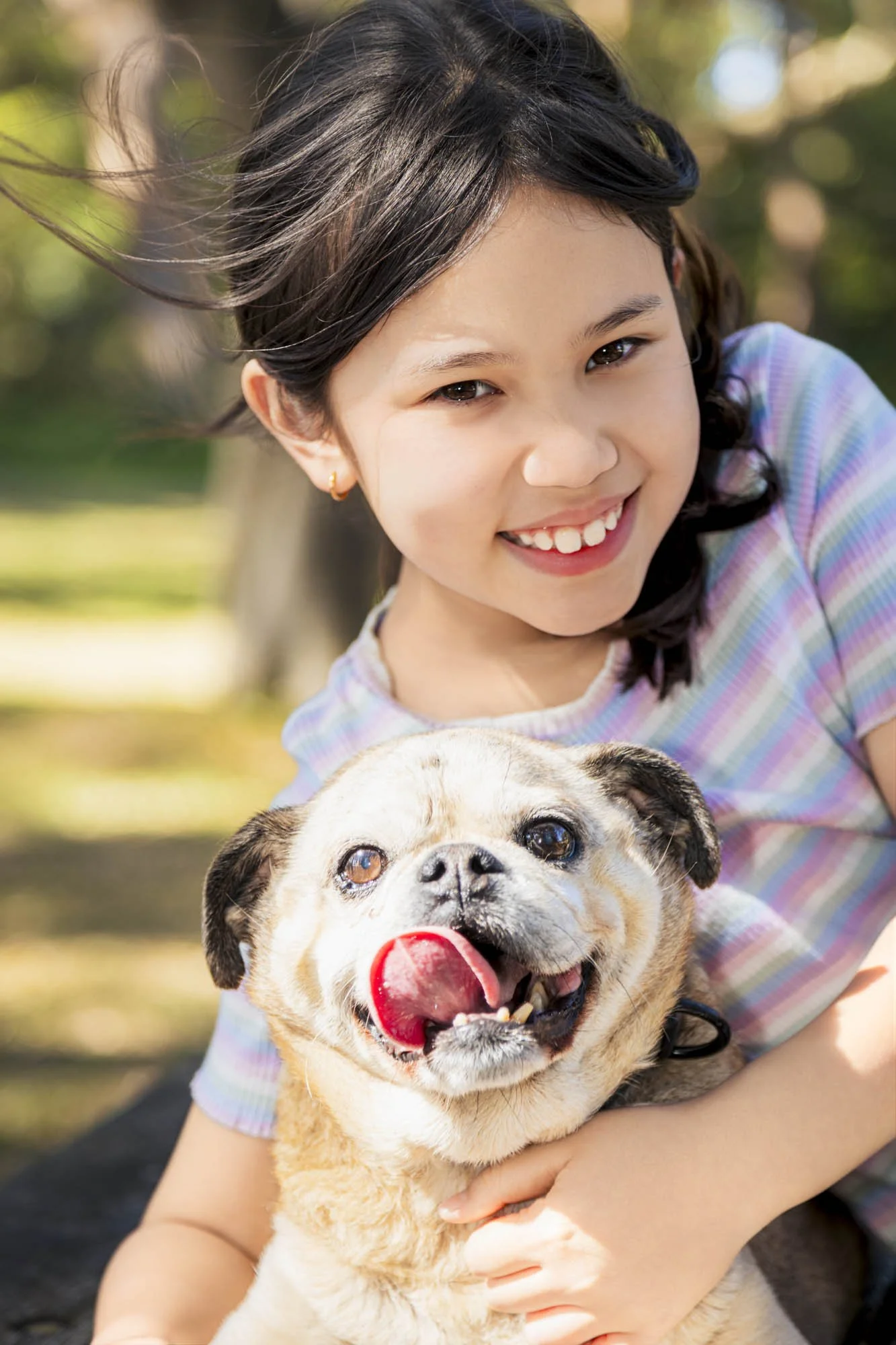 A girl with dark hair smiling and hugging a bulldog with a tongue hanging out outdoors.