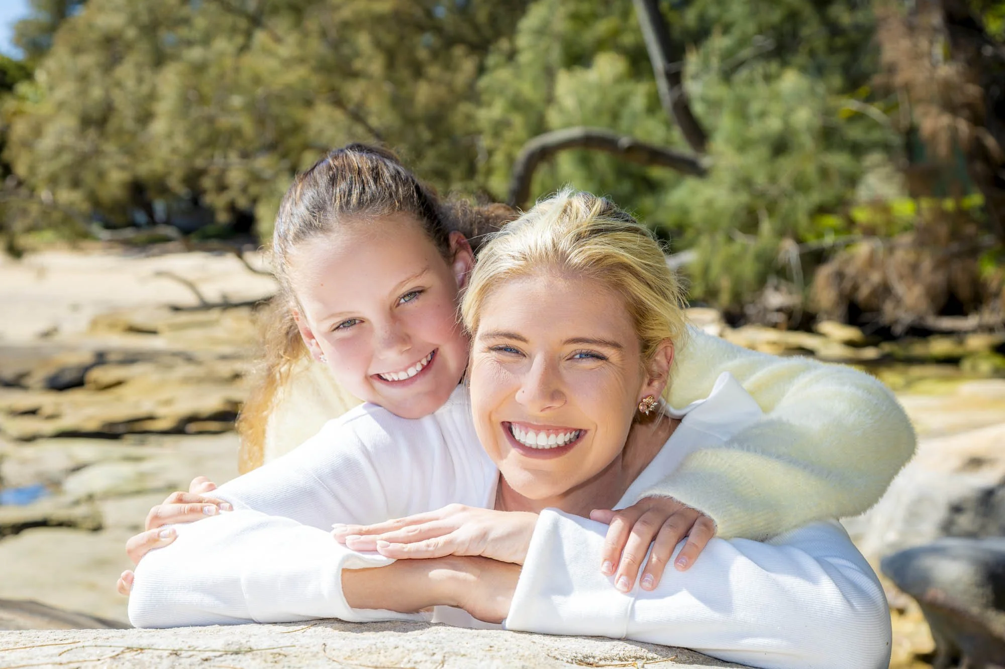 A woman and a girl, likely her daughter, outdoors on a beach, smiling and hugging each other, with trees and sand in the background.