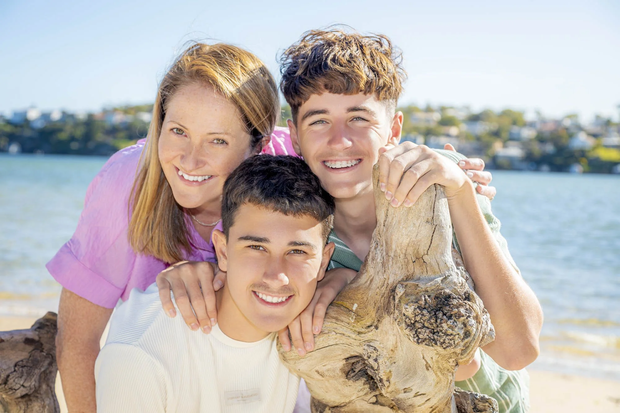 A group of four people, two women and two young men, smiling and embracing each other near a body of water with a cityscape in the background.