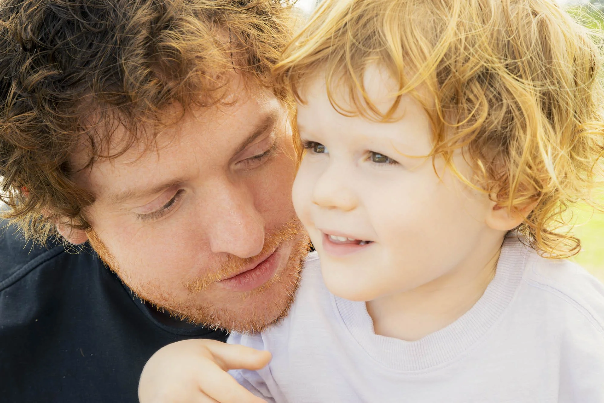 A man with curly brown hair and a beard close to a young girl with curly red hair, both smiling and looking at each other.