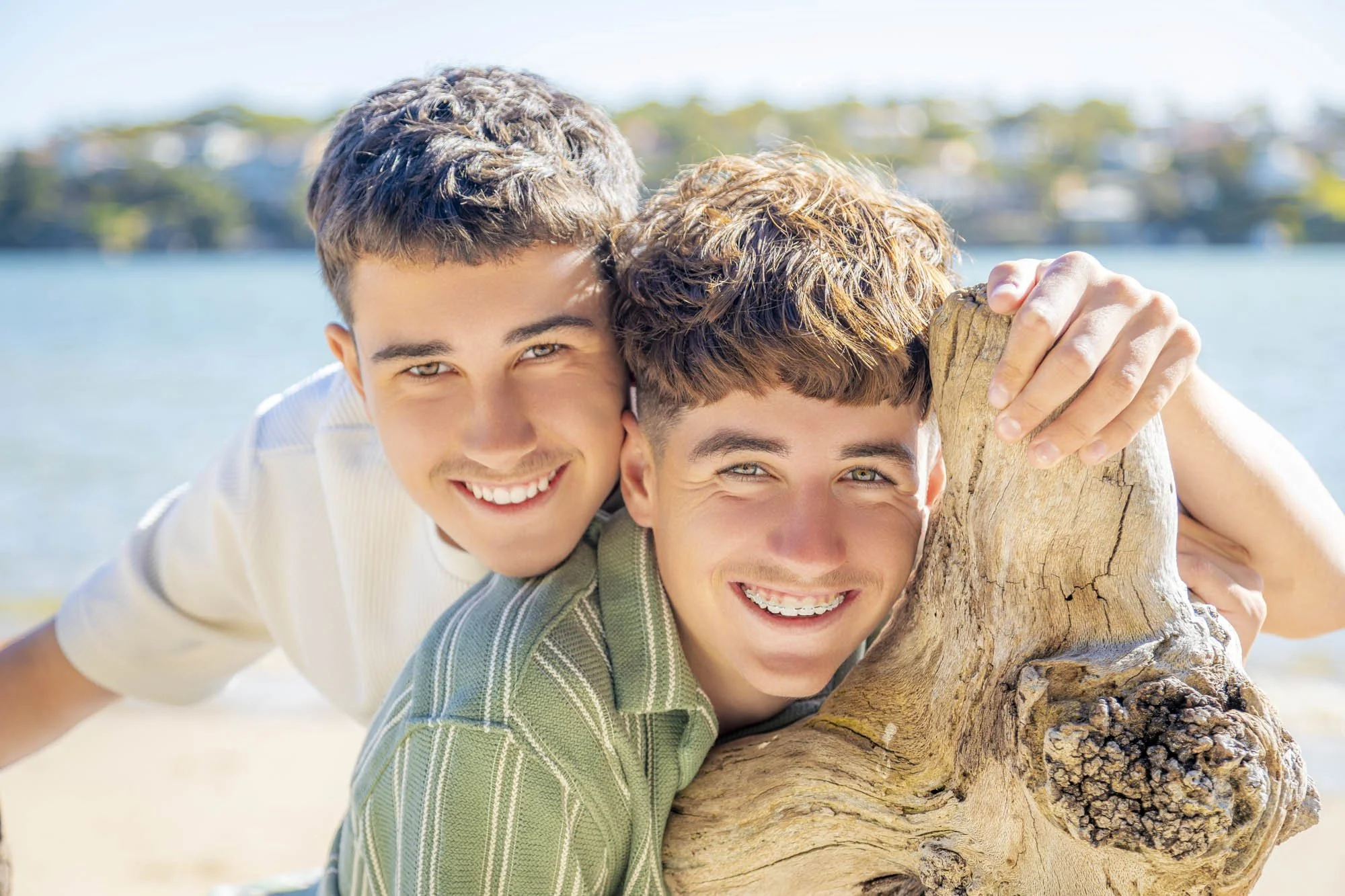Two young men smiling and hugging a large piece of driftwood on a beach with a water body and blurred landscape in the background.