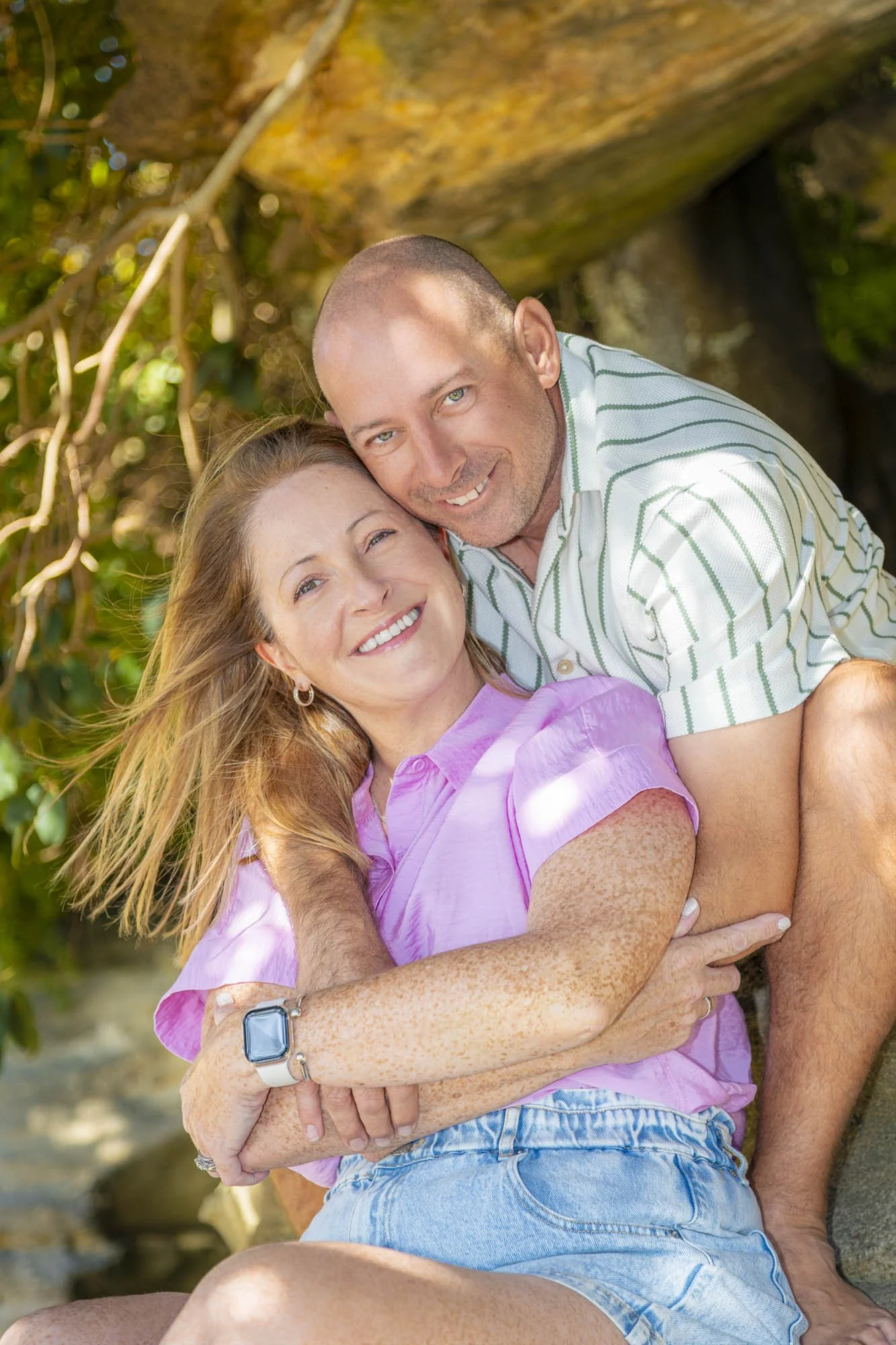 A smiling woman with long red hair and freckles wearing a pink shirt. She is sitting on a person's lap, who is hugging her from behind. The man has short hair, light skin, and is wearing a white and green striped shirt. They are outdoors near a tree 