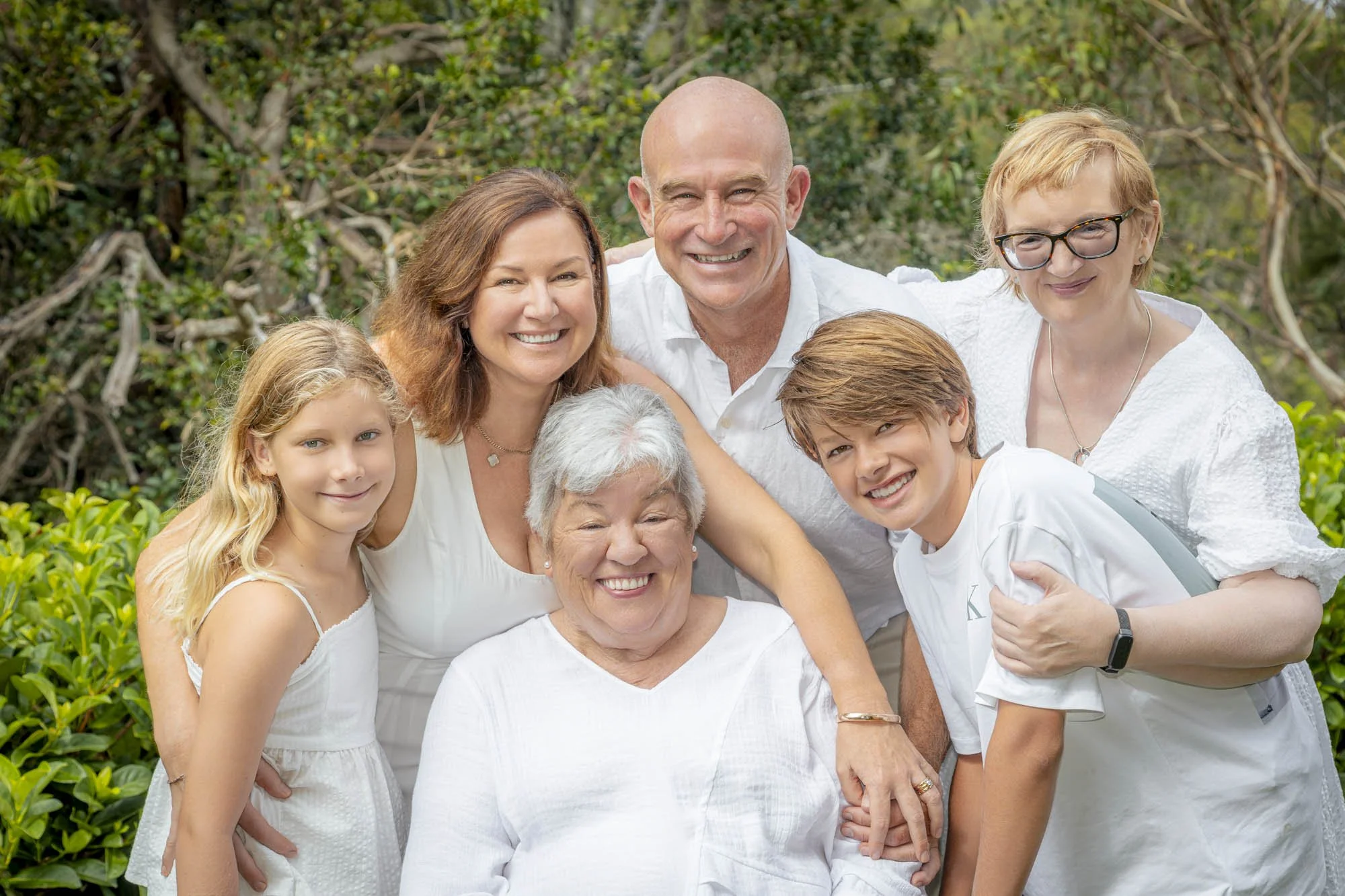 A happy multigenerational family group outdoors, wearing white clothes, smiling, with trees and greenery in the background.
