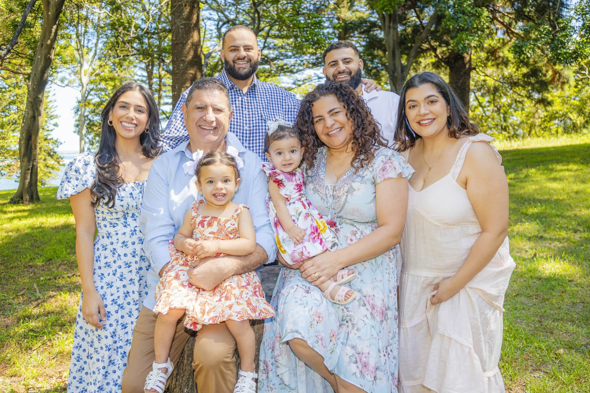 A multi-generational family group portrait outdoors in a park with green trees in background. The family includes women, men, and children smiling and sitting together on a bench, wearing casual summery clothing.