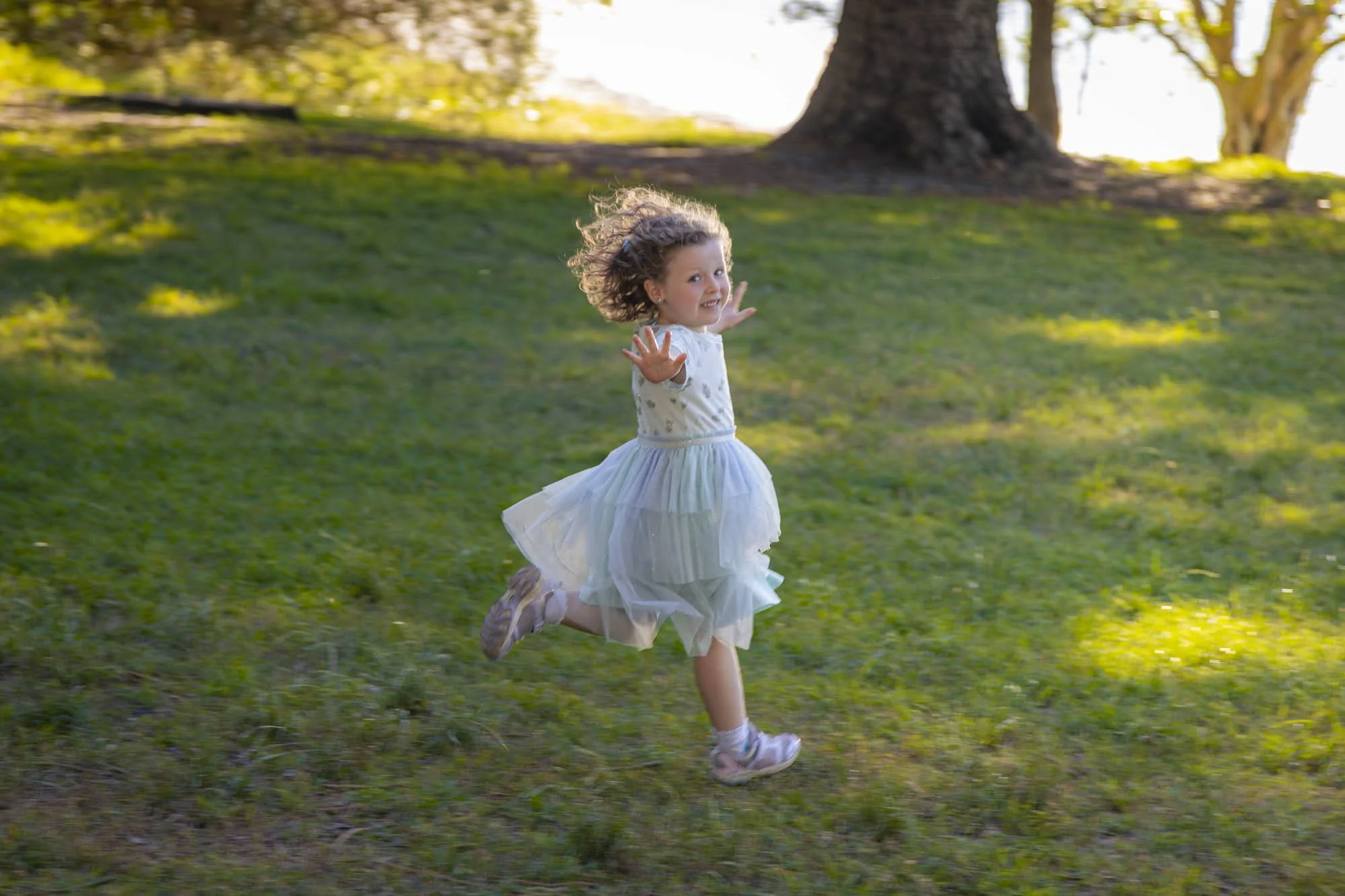 A young girl with curly hair, wearing a white dress with sparkles, running on a grassy field with a large tree in the background and sunlight filtering through the trees.