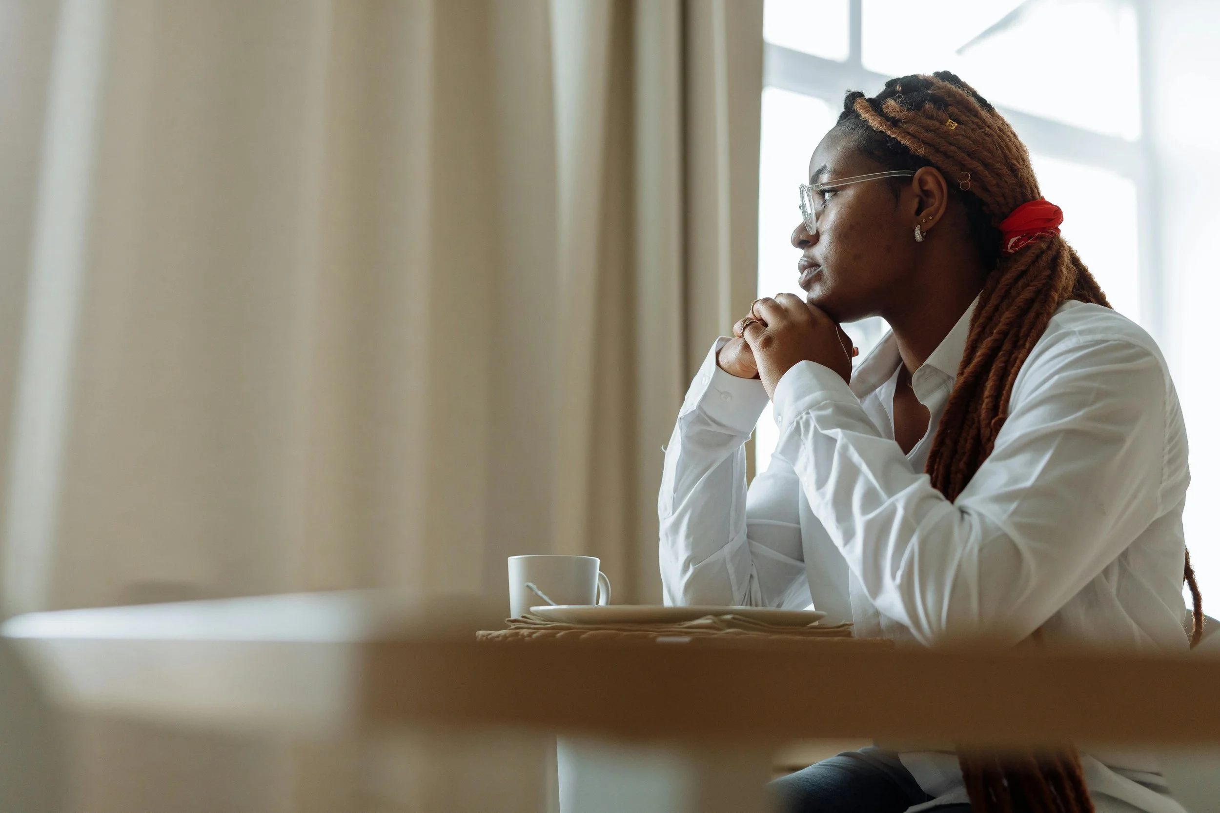 Black woman with locs sitting at a table in quiet thought, looking toward a window
