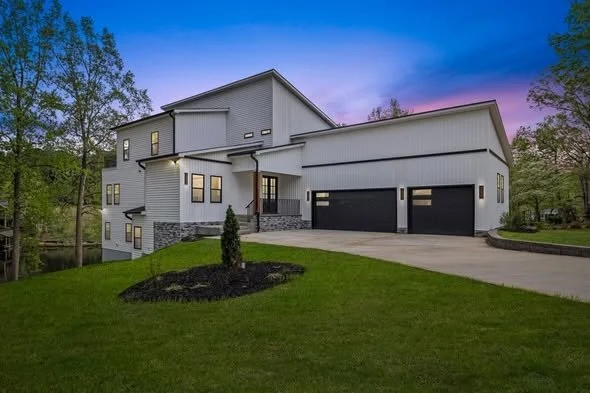 Modern white house with multiple levels, large garage, and surrounding greenery at dusk.