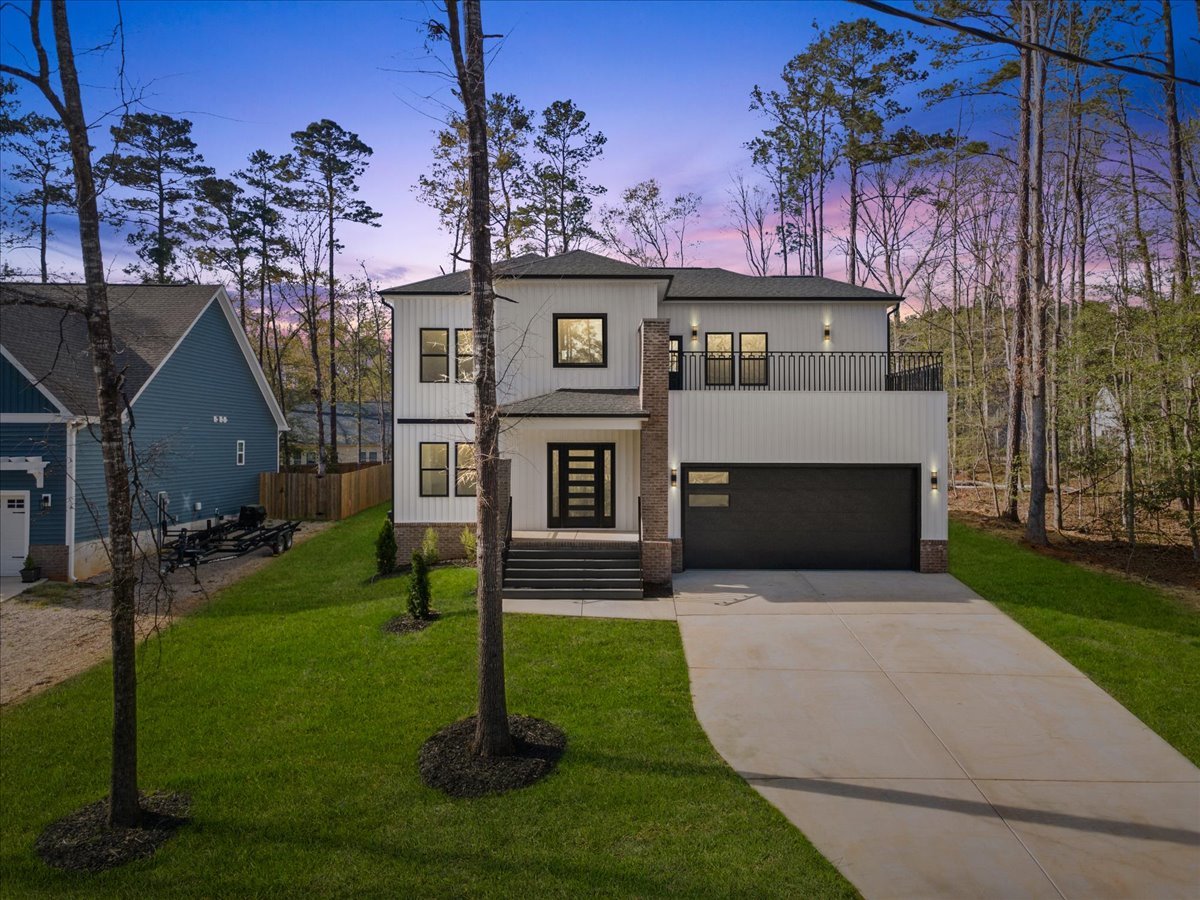 Modern two-story house with a white exterior, black accents, and a flat roof, featuring a small front porch, a driveway, and a balcony on the upper level. The house is surrounded by a grassy yard with trees, with a sunset sky in the background.
